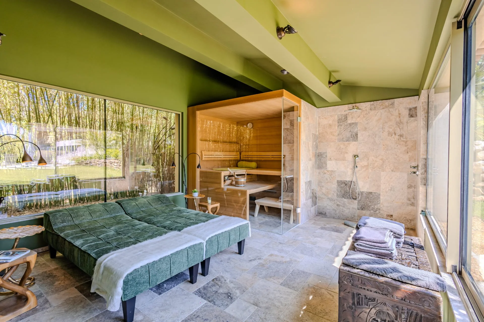 A spacious indoor wellness or spa room with large windows showing a bamboo garden outside, featuring a green tufted lounge bed, a wooden sauna, a walk-in shower area with stone tile walls, and lined towels on a bench.