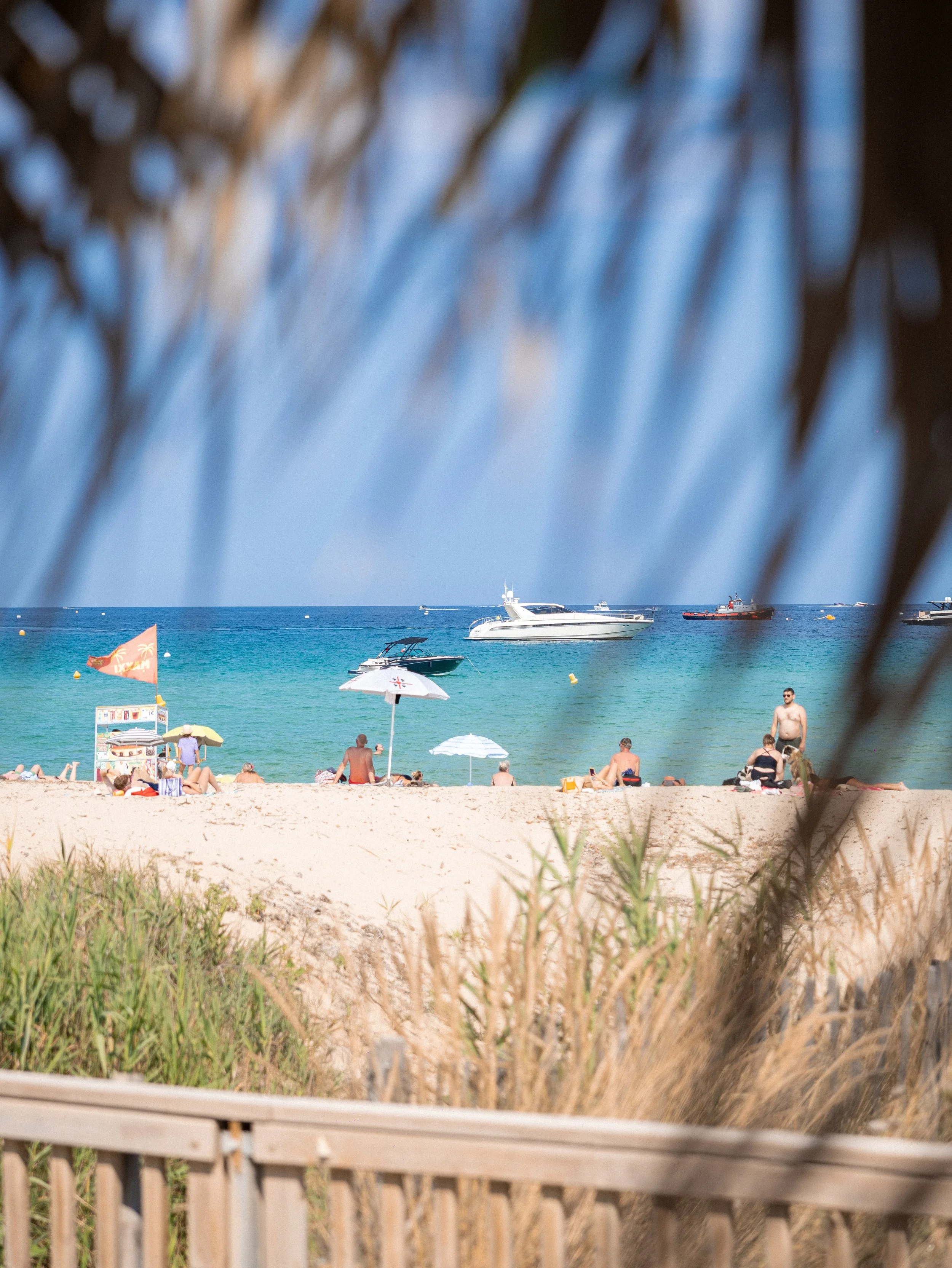 Beach scene with people relaxing on the sand, umbrellas, and boats on the water, seen through a wooden fence and some greenery in the foreground.