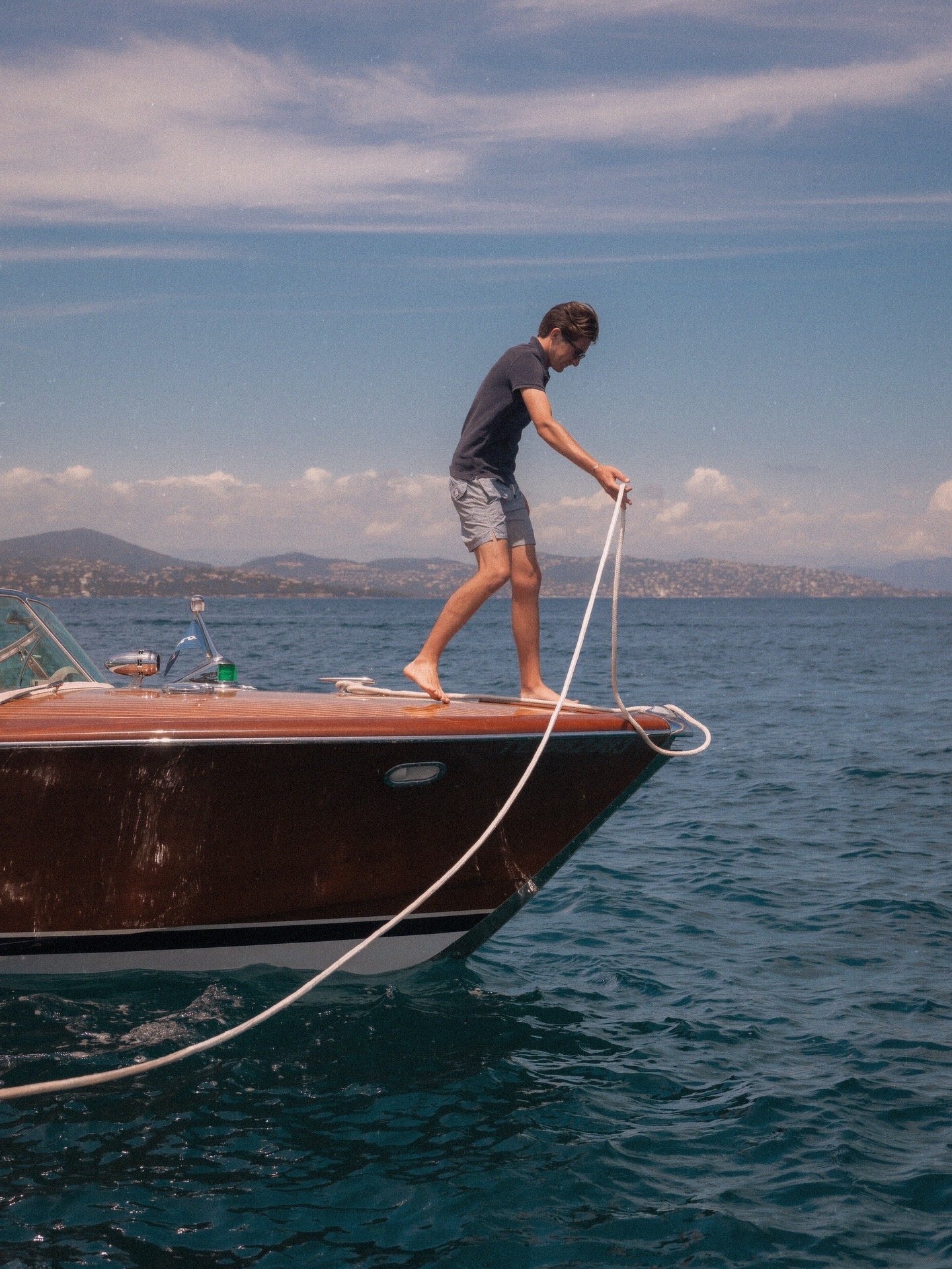 A man standing barefoot on the front of a boat, tying a rope, with water and a distant shoreline in the background.