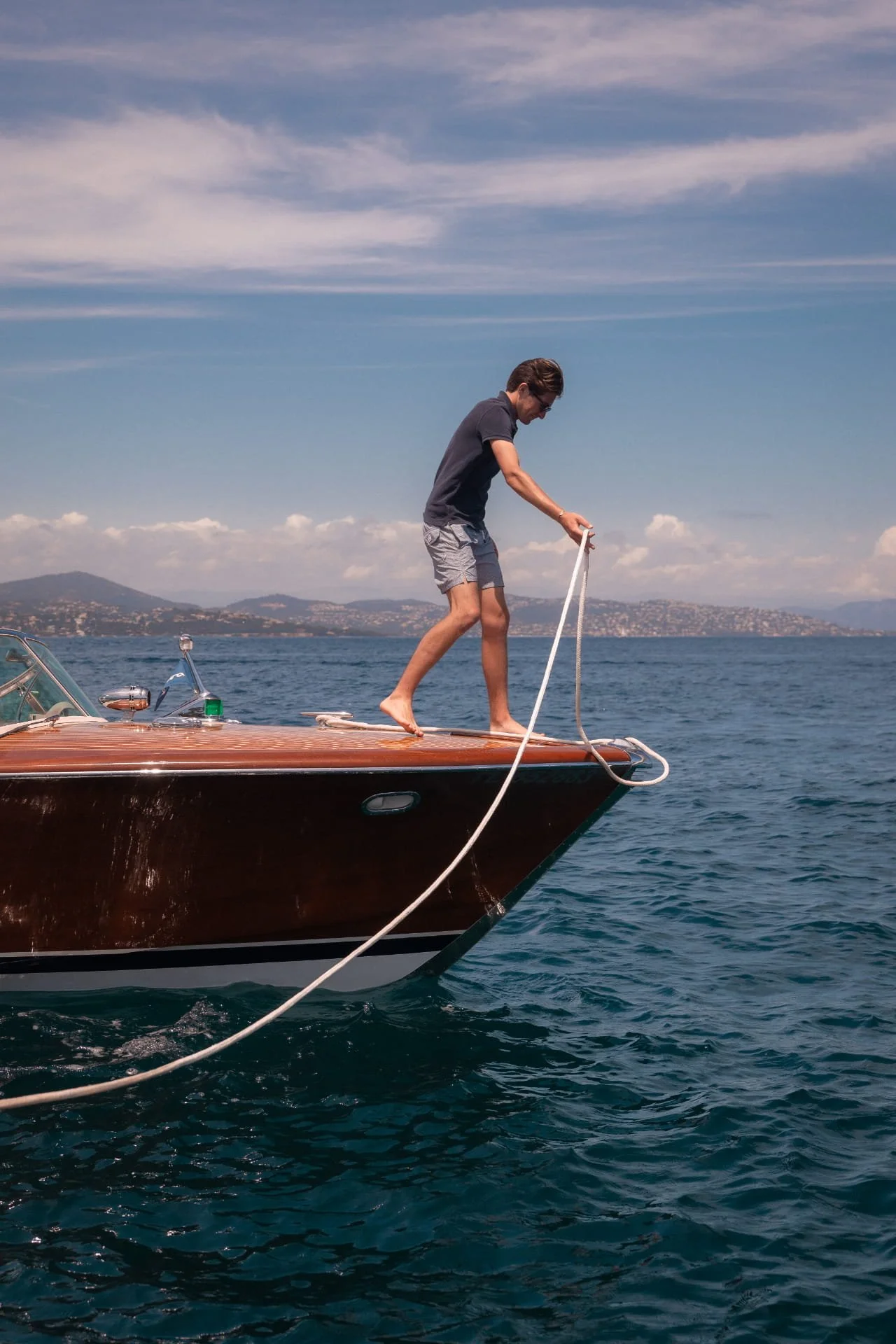 A man in a dark t-shirt and shorts standing on the bow of a wooden boat, holding a rope while on the water during daytime with a distant land and sky with clouds.