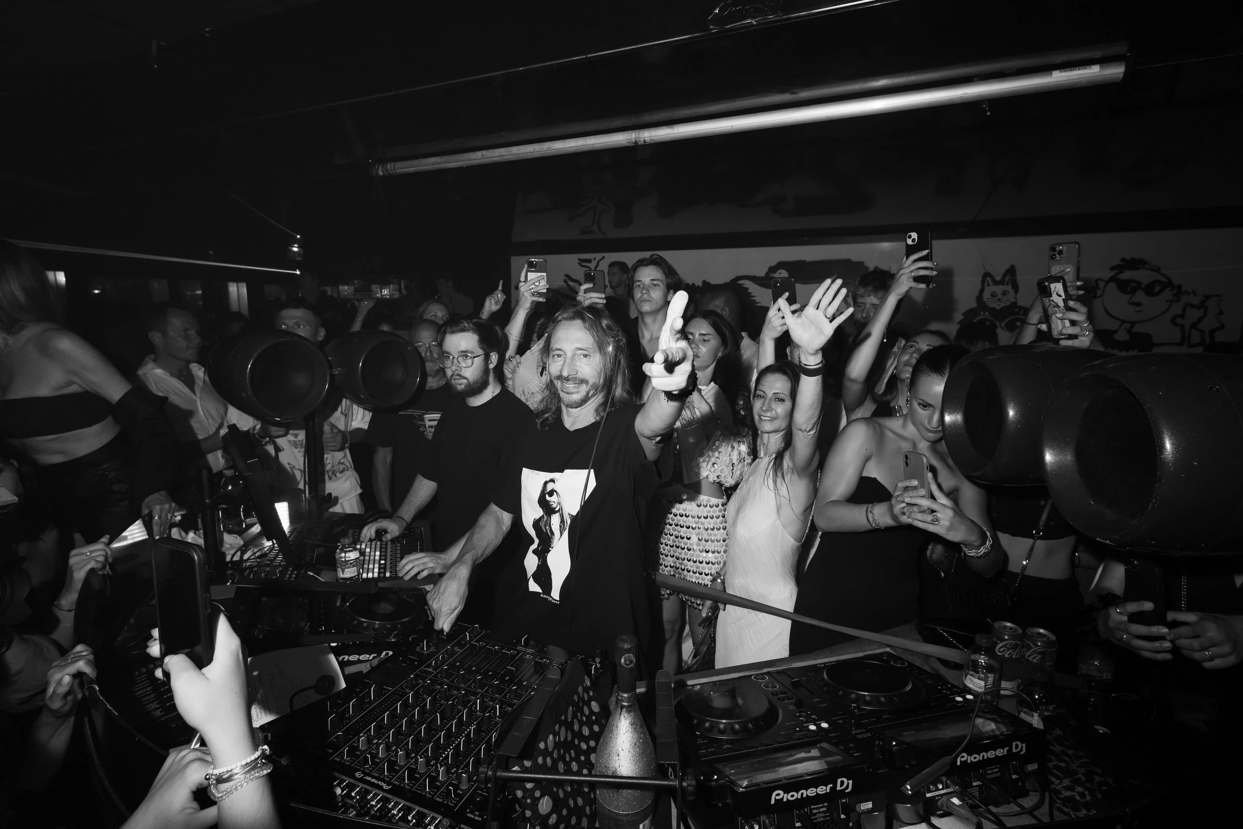Crowd of people dancing and taking photos at a DJ concert in a dark indoor venue, with DJ equipment visible in the foreground.