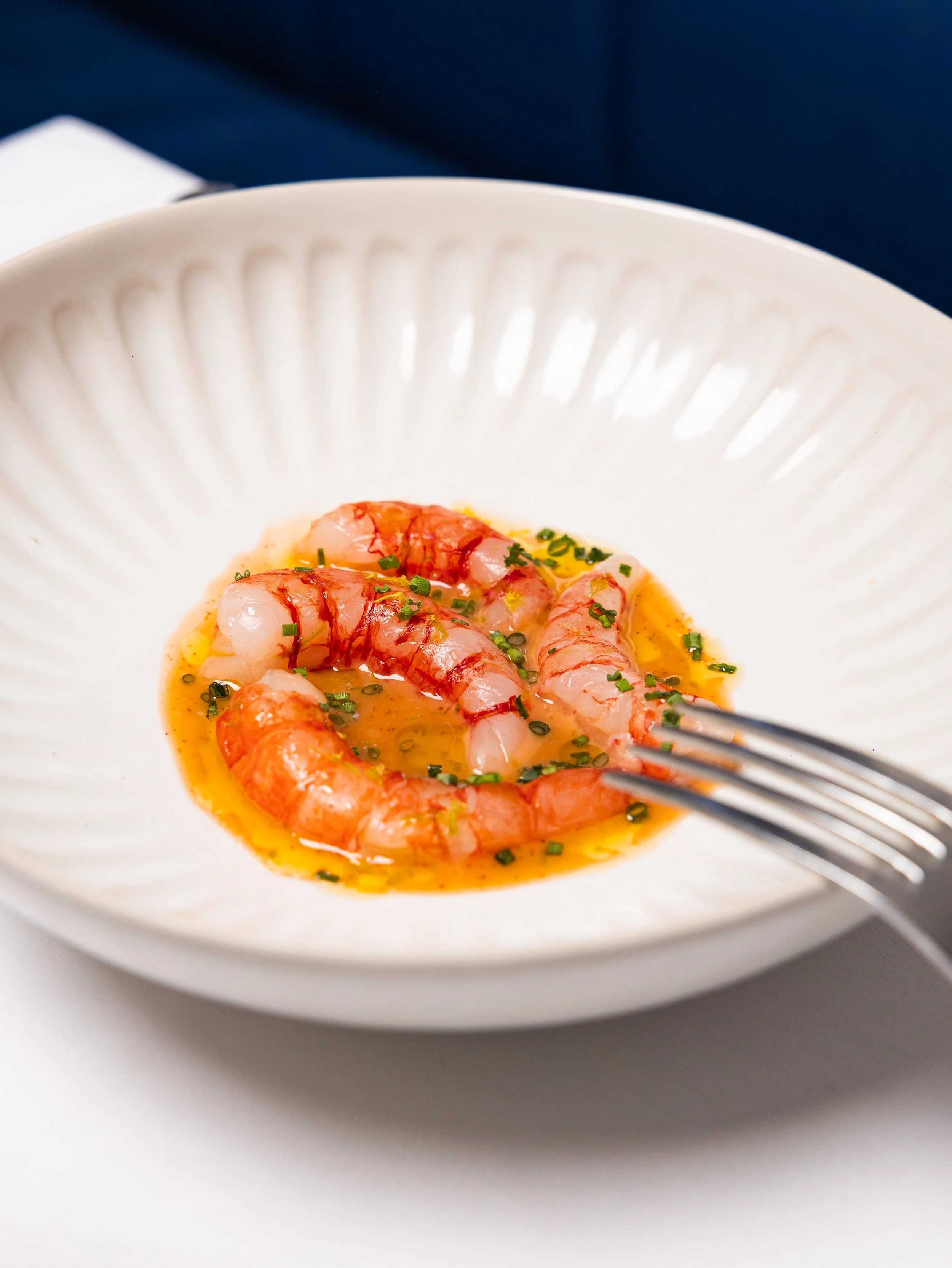 Close-up of cooked shrimp in a white bowl with garnishes and sauce, on a white surface with a blue background.