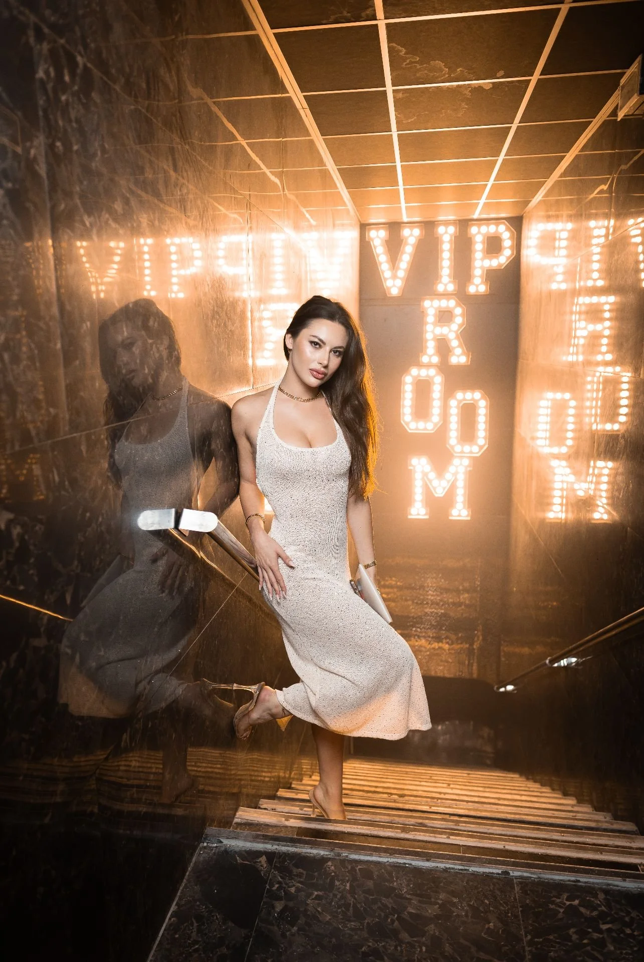 A woman in a white dress and high heels is sitting on stairs in an elevator with reflective black marble walls. Behind her, there is a large illuminated sign with the words 'VIP ROOM' in bright lights.
