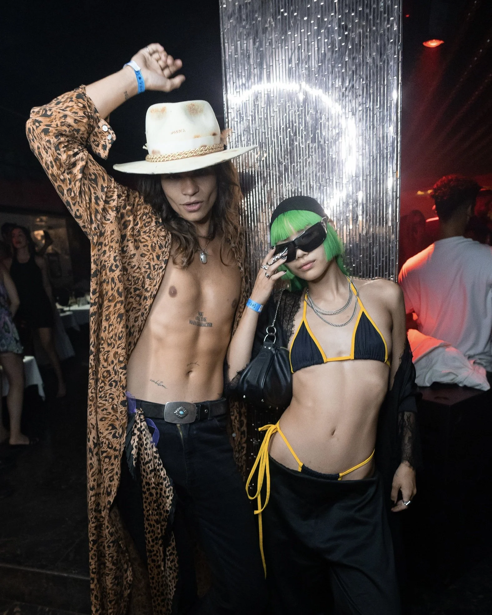 Two individuals in a nightclub or party setting. The person on the left is shirtless, wearing a leopard print jacket and a wide-brimmed white hat with a gold chain detail. The person on the right has bright green hair, is wearing black sunglasses, a 