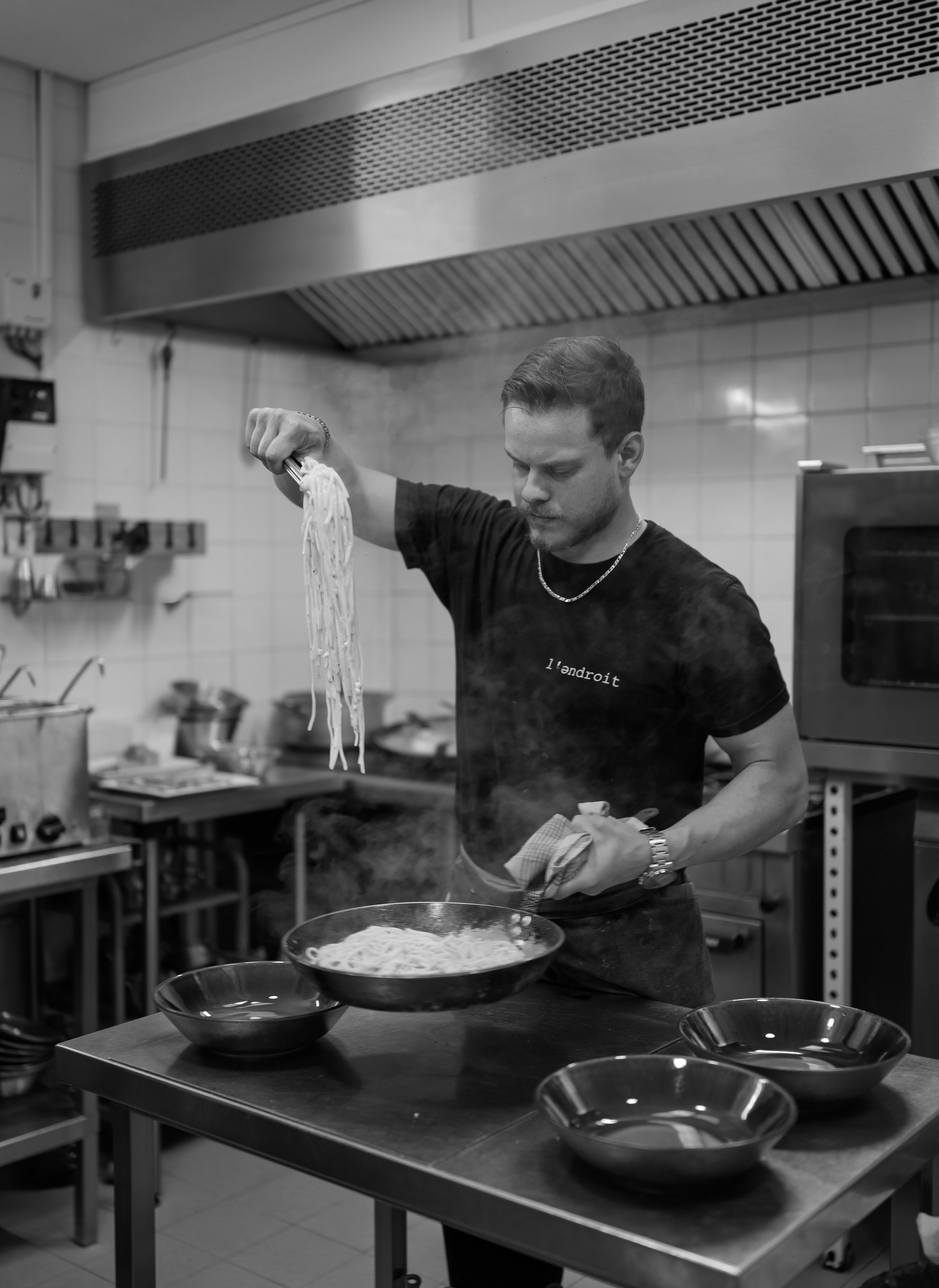 A man preparing spaghetti in a commercial kitchen, holding a fork with noodles and a bowl of cooked pasta on the counter.