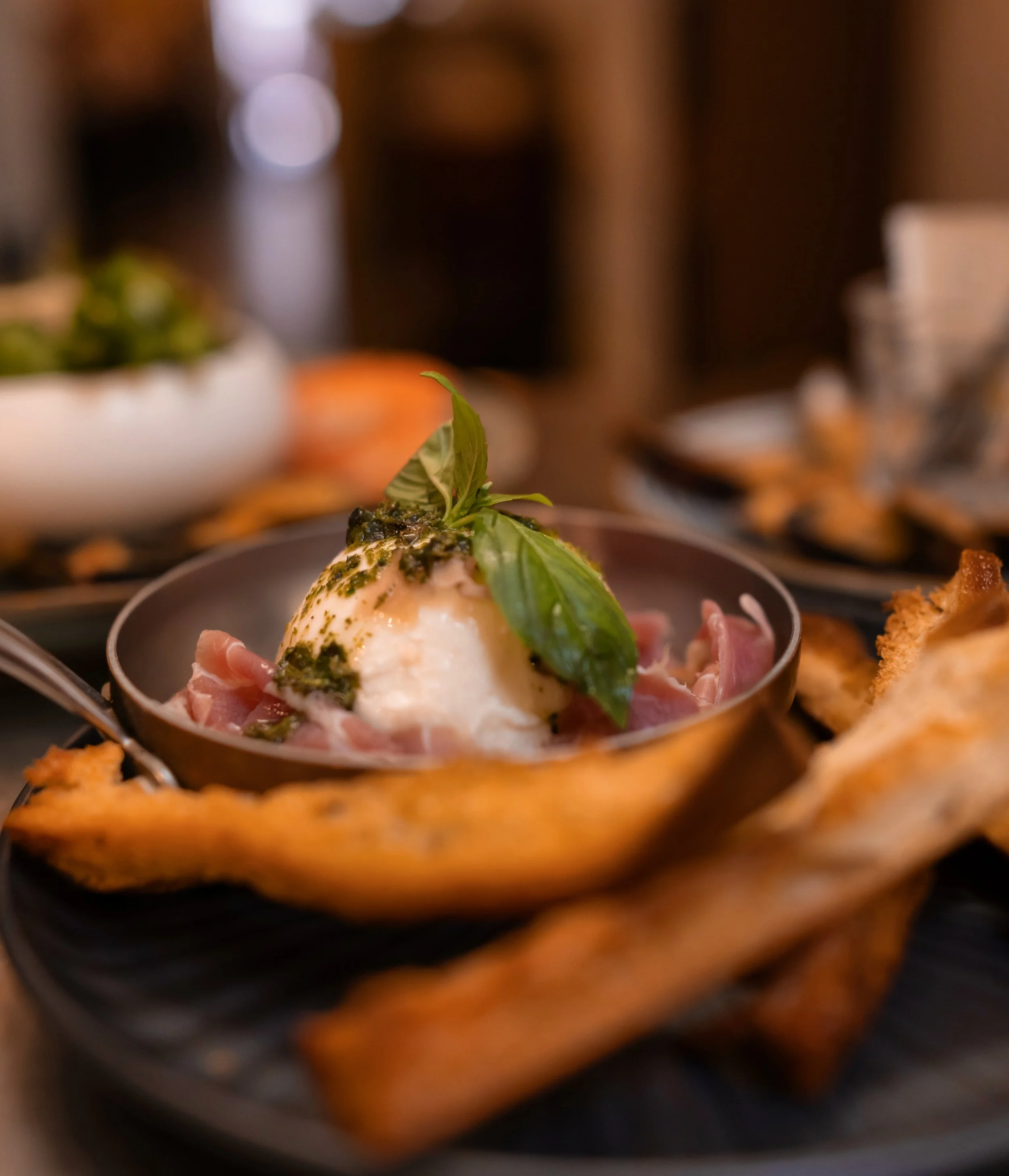 A close-up of a dish with a soft-boiled egg topped with pesto, basil leaves, and herbs, served with cured meat, on a black plate. There are other food items and a blurred background.