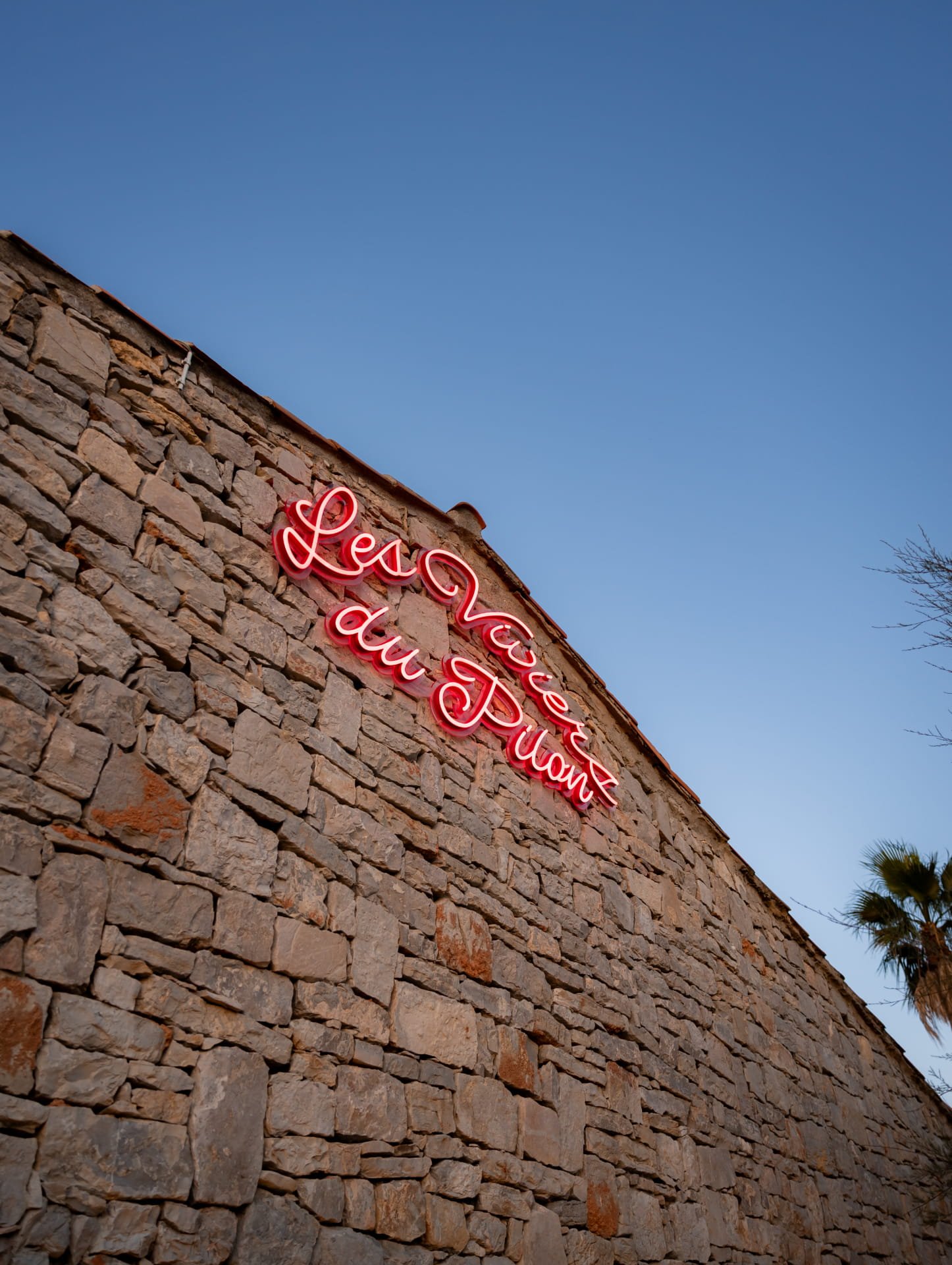 A stone building with a red neon sign reading 'Love' on it, against a clear blue sky, with a tree branch visible on the right edge.