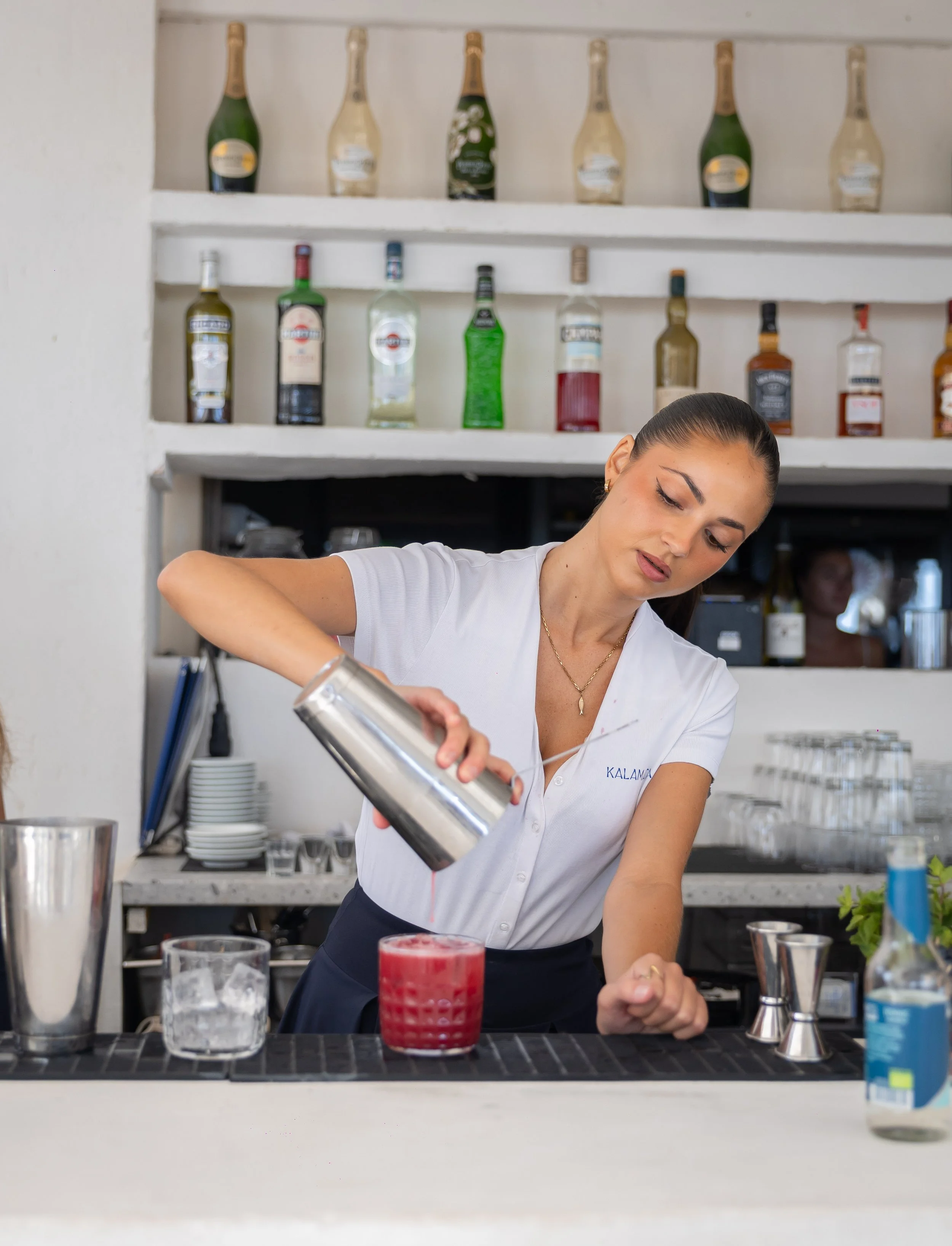 A woman with dark hair tied back, wearing a white shirt and a necklace, is pouring a drink into a glass at a bar or restaurant counter. There are various bottles on shelves behind her, including bottles of sparkling wine and liquor. Several glasses, 