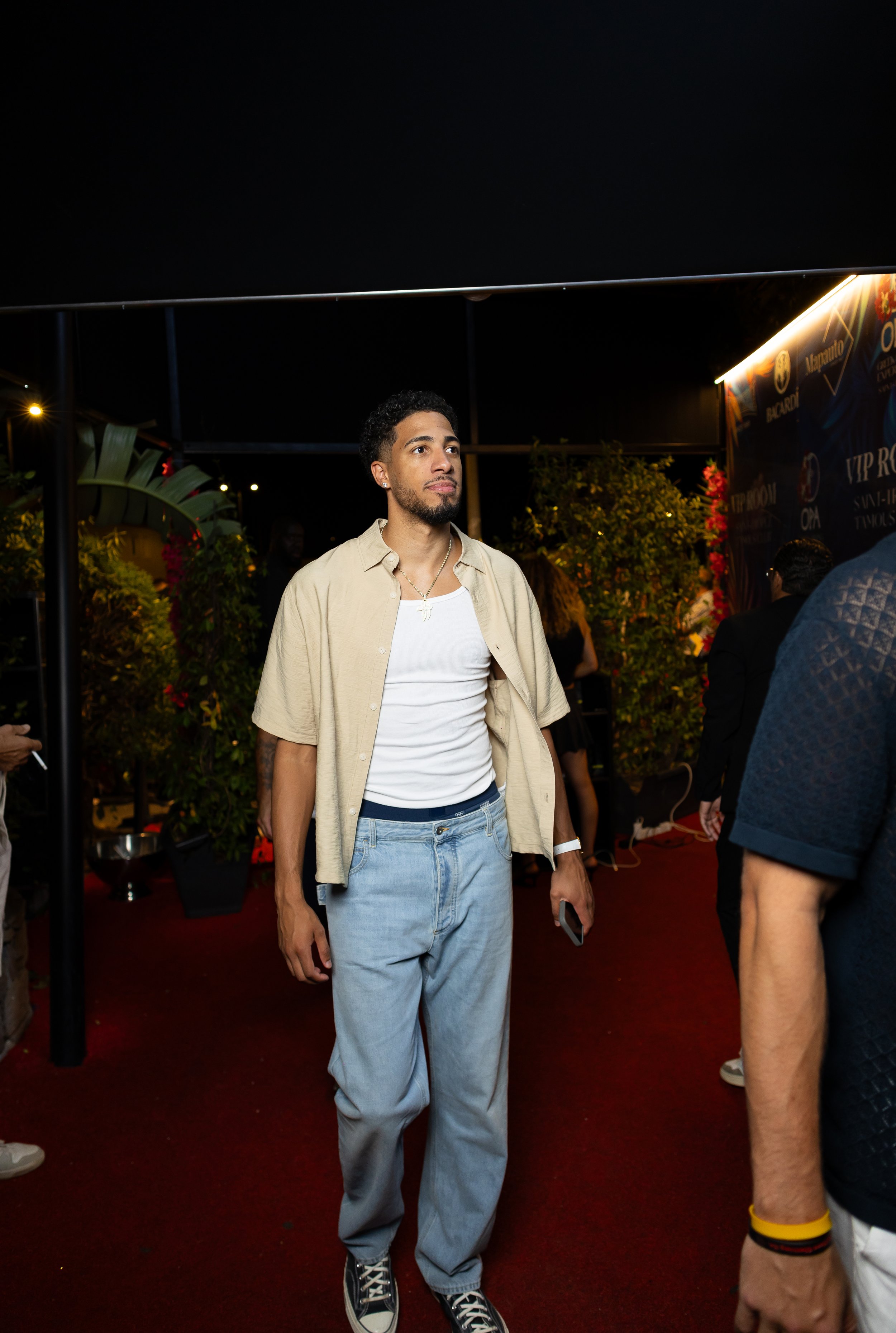 Young man with curly dark hair, light beard, wearing a beige open shirt over a white t-shirt, light blue baggy jeans, and black high-top sneakers, standing on a red carpet at night surrounded by other people, with outdoor plants and a brightly lit si