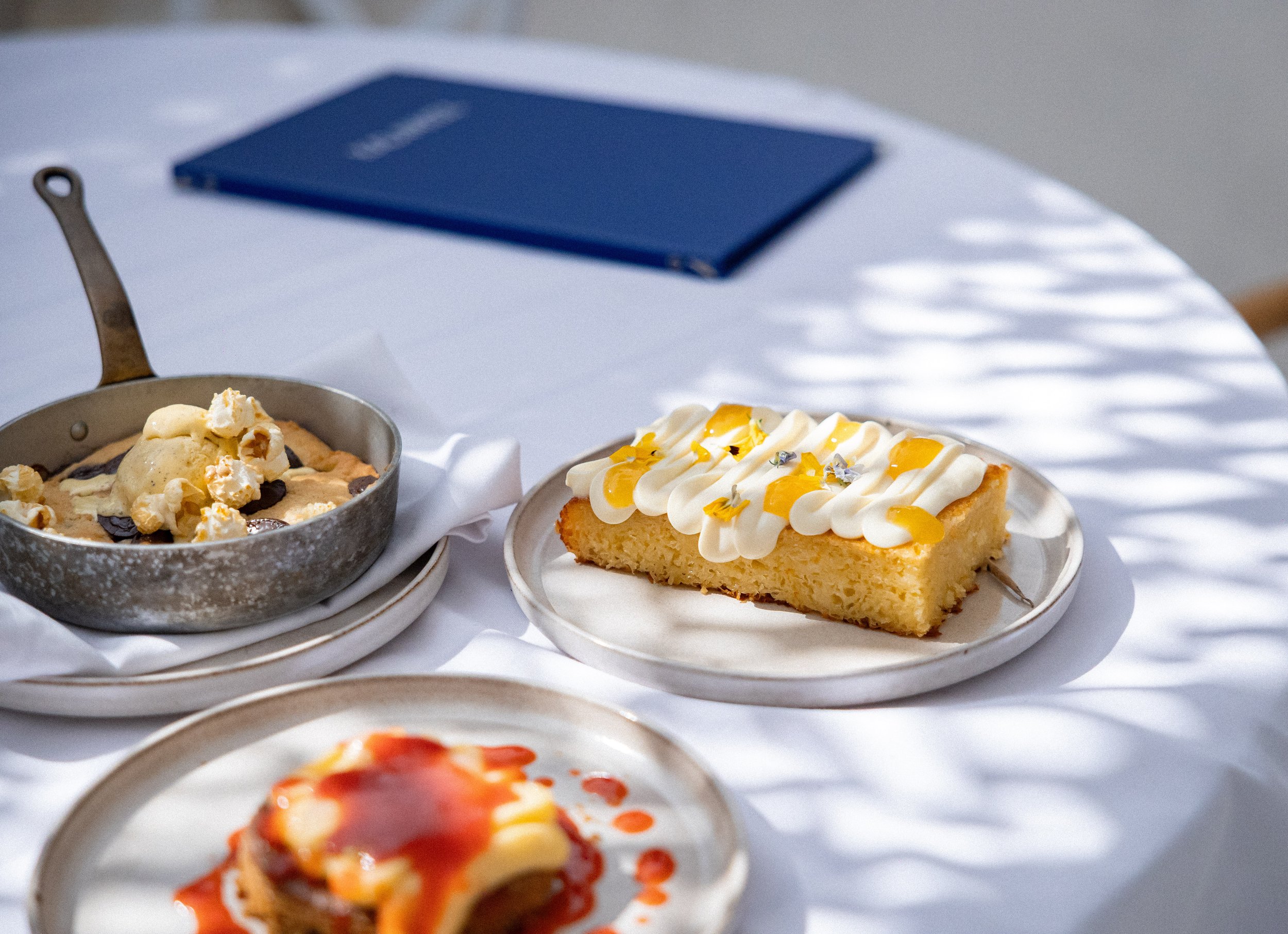 Close-up of assorted desserts on plates, including a slice of cake with icing and edible decorations, and a dessert topped with whipped cream and popcorn, with additional dishes in the background on a white tablecloth.