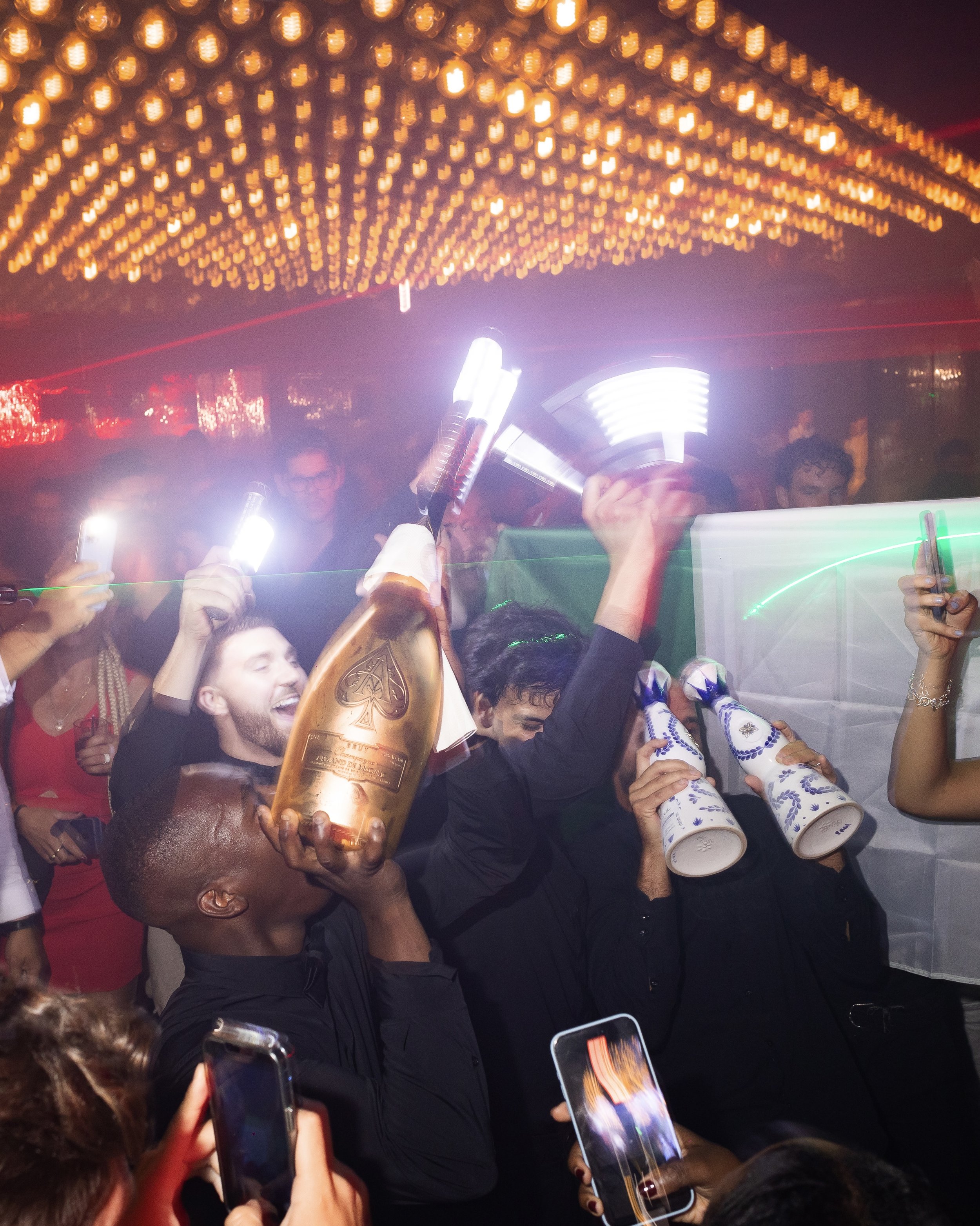 A crowded party scene with people holding smartphones and taking photos or videos. There is a person holding a large bottle of Ace of Spades champagne. The ceiling has many small, warm-colored lights. The atmosphere is lively and celebratory.