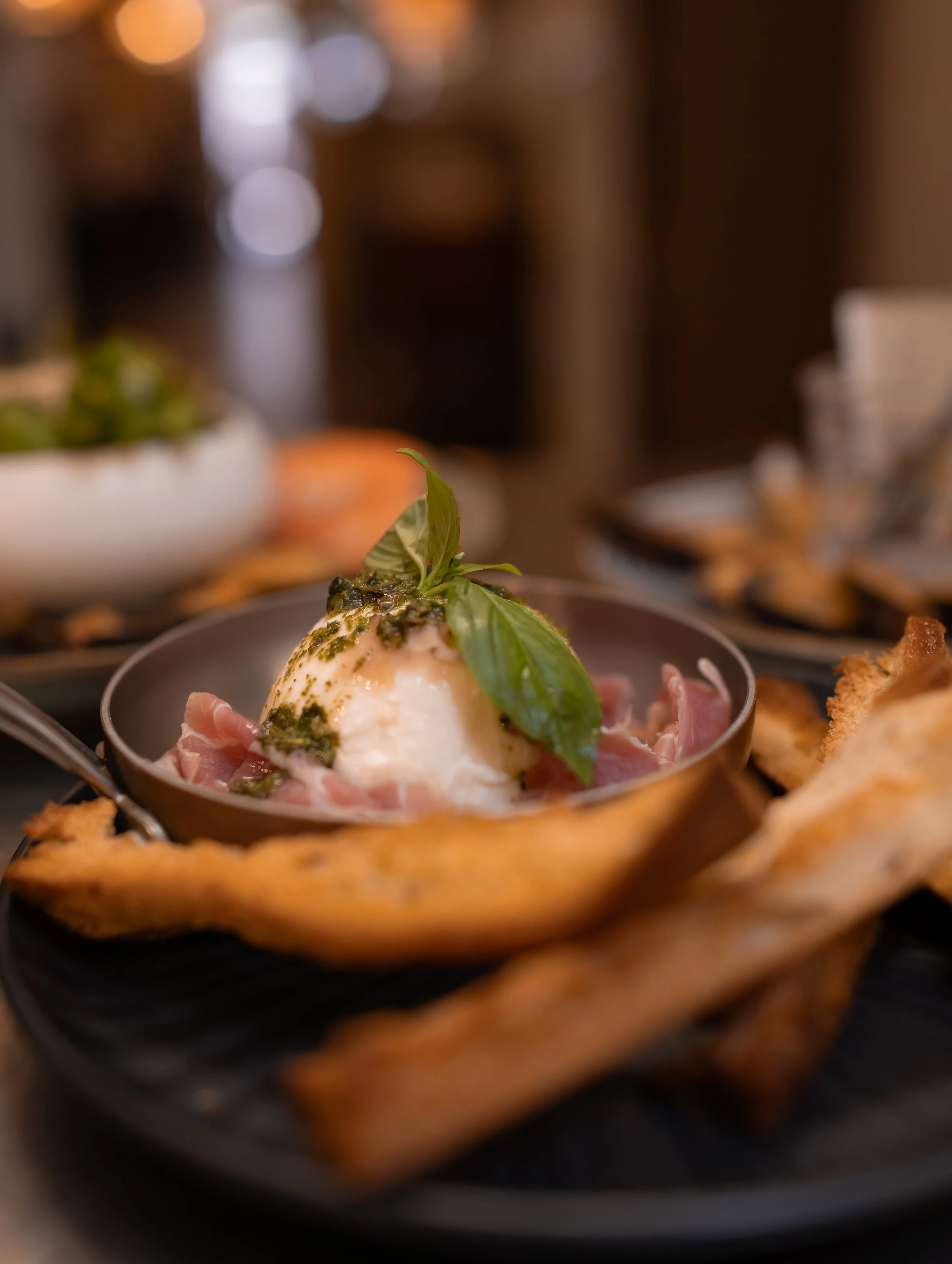 Close-up of a poached egg topped with basil and pesto, served in a small metal dish, with other dishes blurred in the background on a dining table.