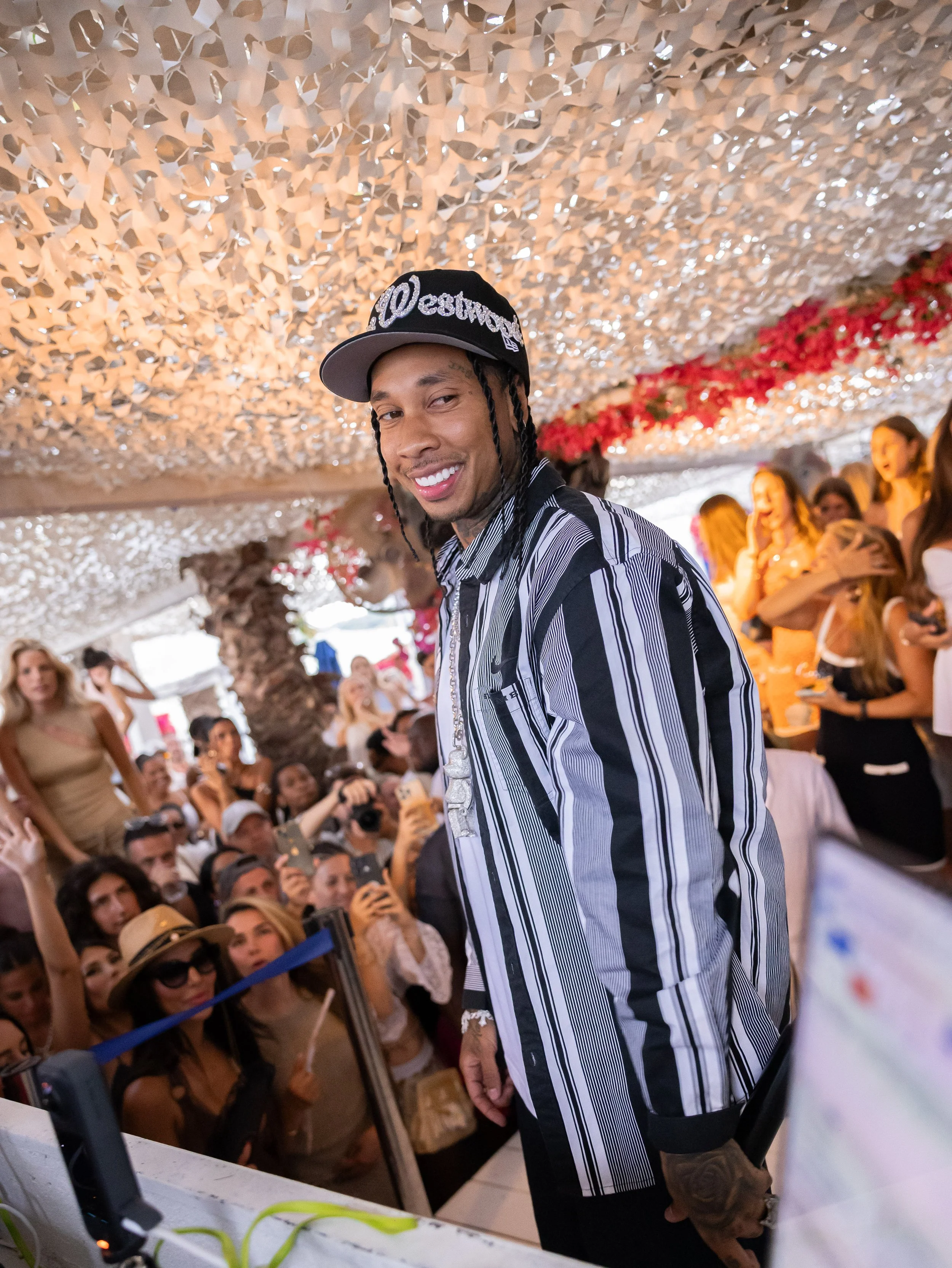 A man with braided hair wearing a black baseball cap and a striped shirt, smiling at the camera at an indoor event with a decorated ceiling and a crowd of people in the background.