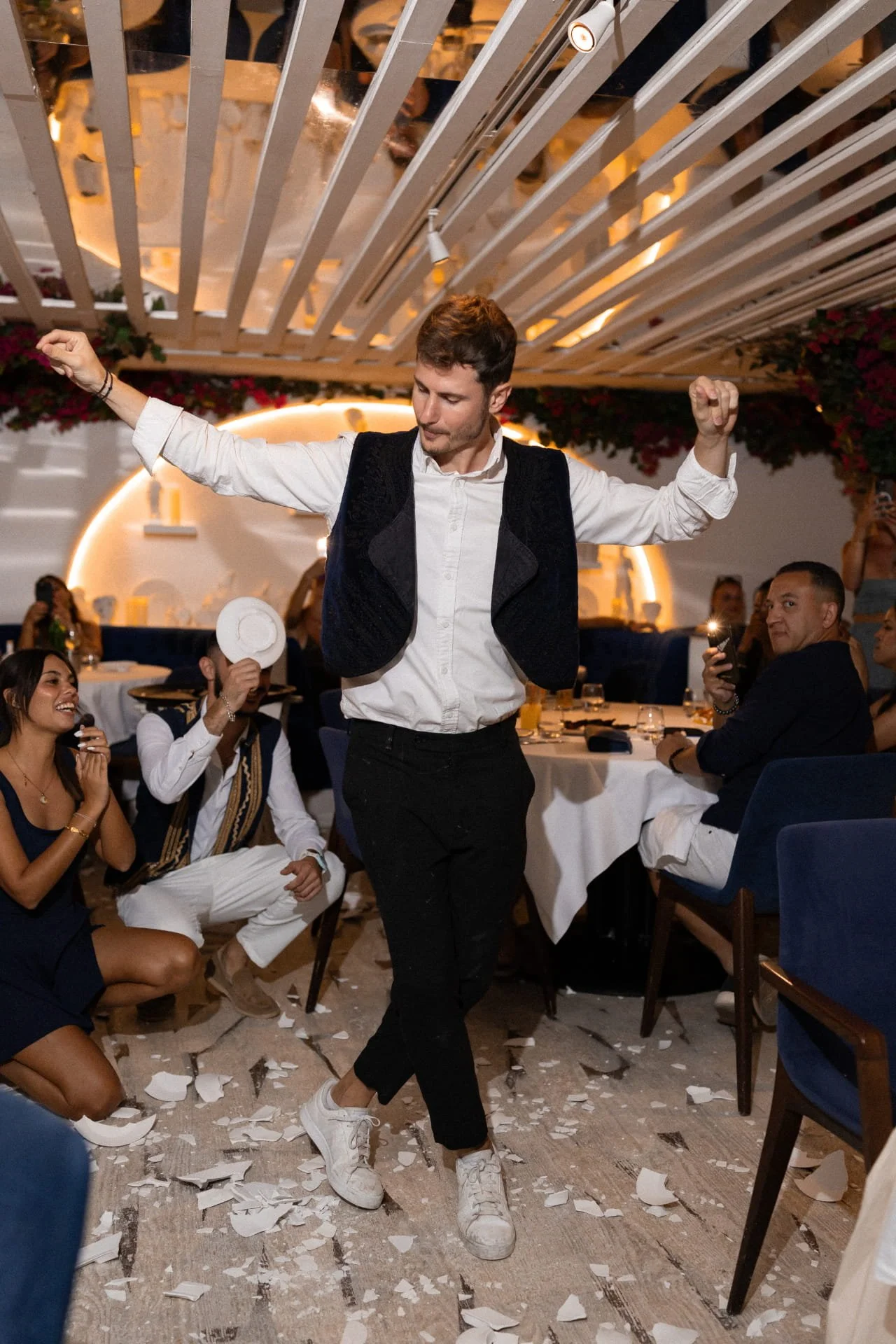 A man dancing at a celebration with people seated around tables, some taking photos, and paper confetti on the floor.