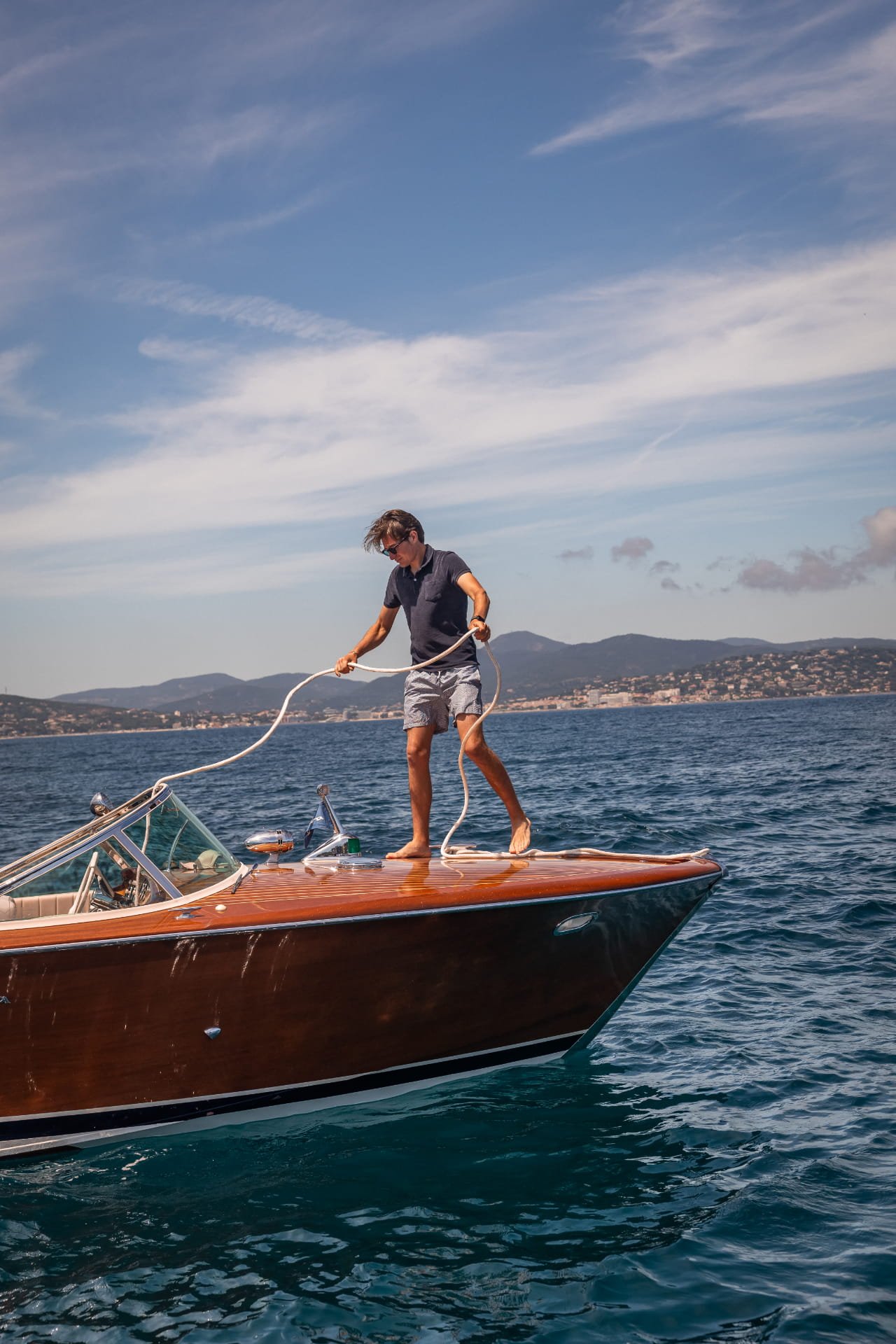 A man standing on a boat holding a rope, with water and distant land in the background on a sunny day.