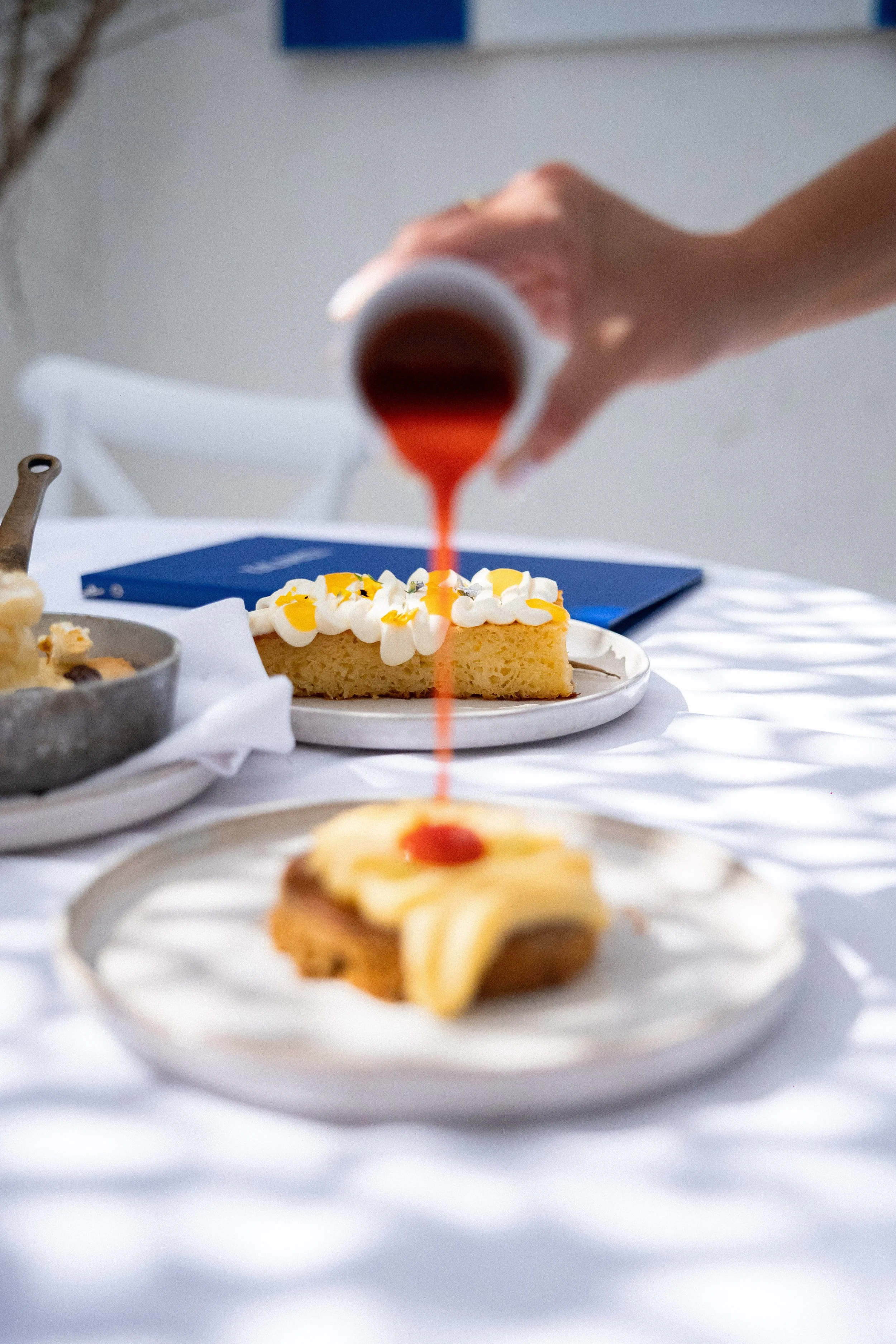 A hand pouring red sauce over a slice of cake on a white plate. Other desserts are partially visible on the table, which is covered with a white tablecloth.