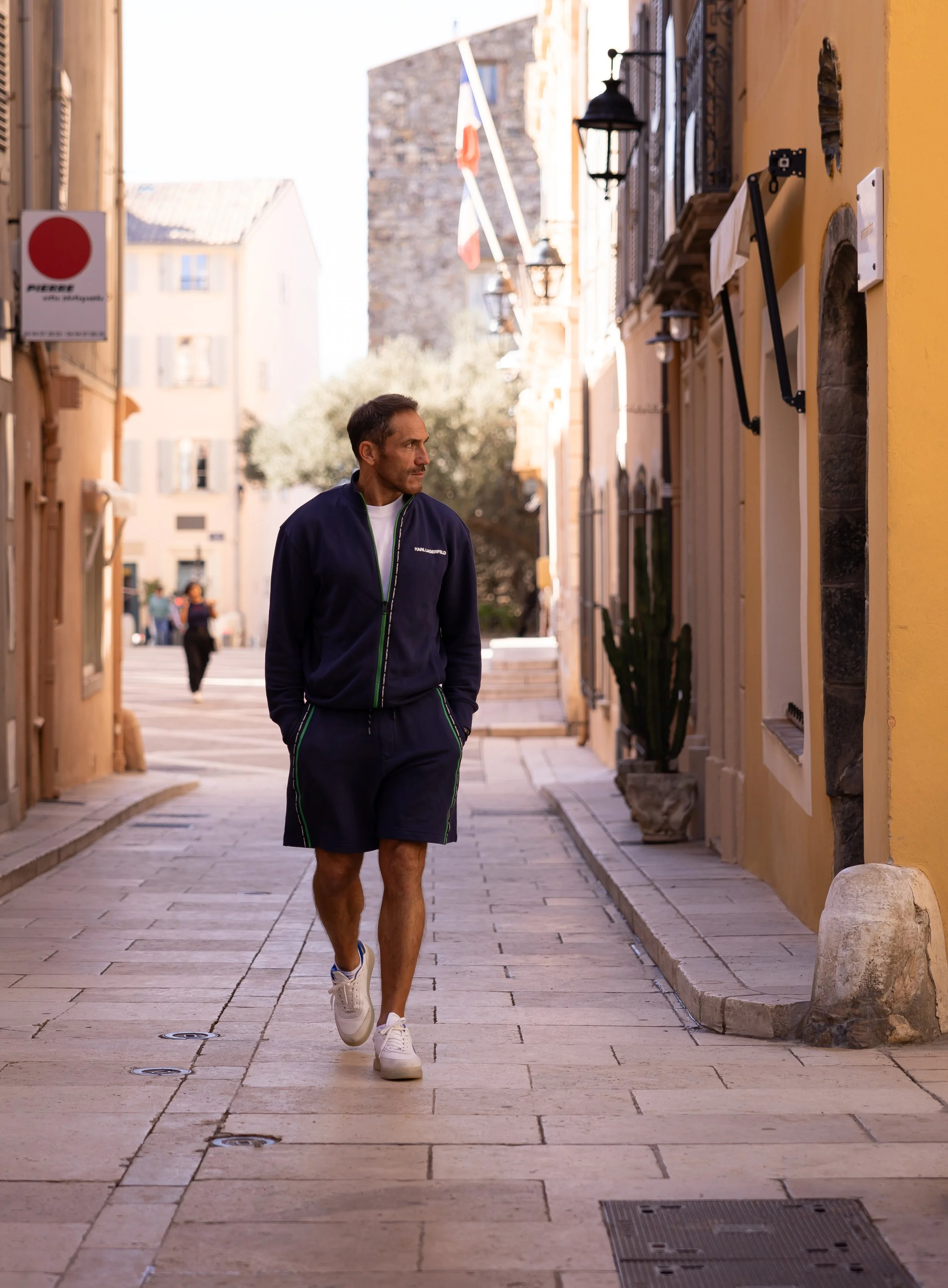 A man walking on a cobblestone street in a European town, wearing a navy jacket, black shorts, and white sneakers, with historic buildings and flags in the background.