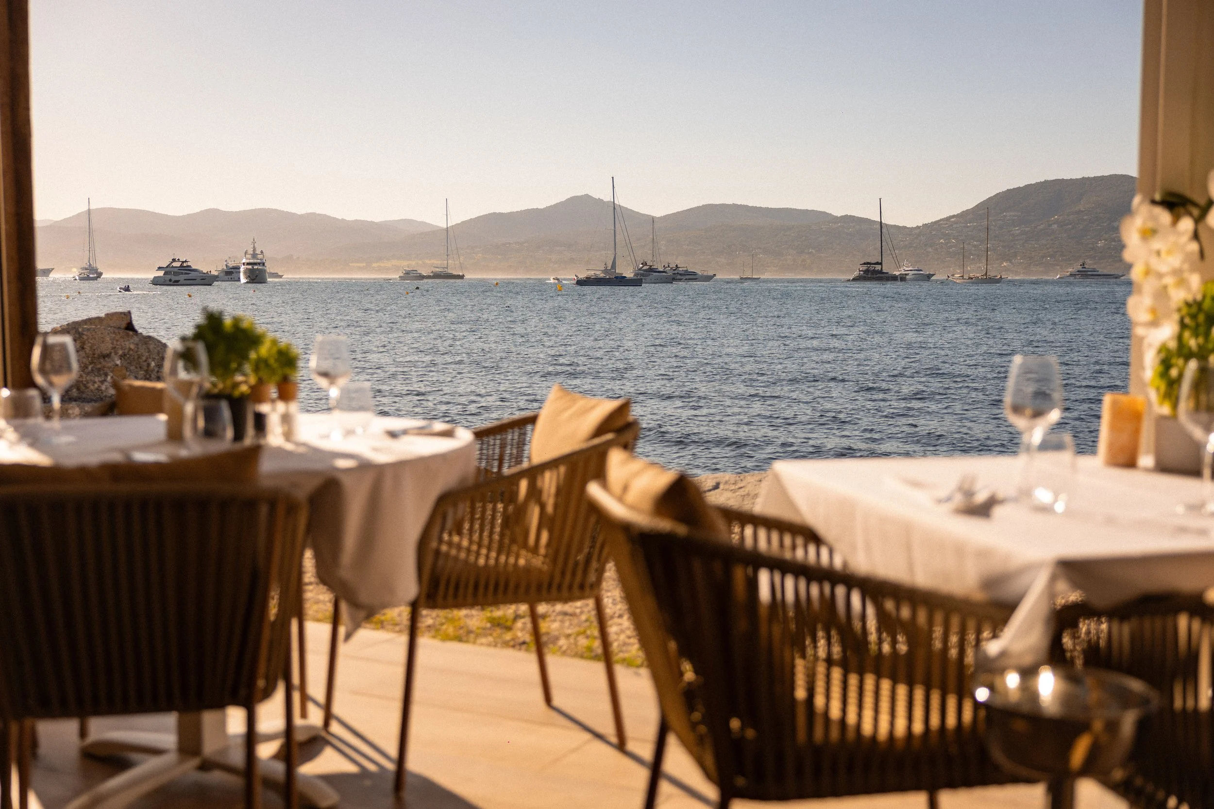 Dining tables set with glasses and flowers overlooking a bay with sailboats and hills in the background