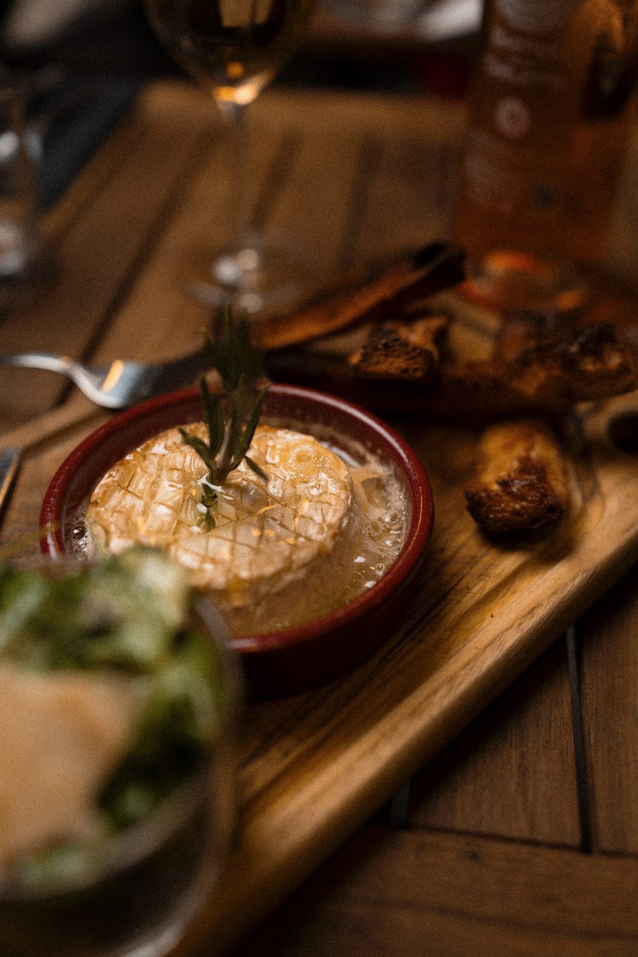 A wooden serving board with grilled meats and a small bowl of creamy dip topped with a sprig of rosemary. The background includes a blurry glass of beer and other condiments.