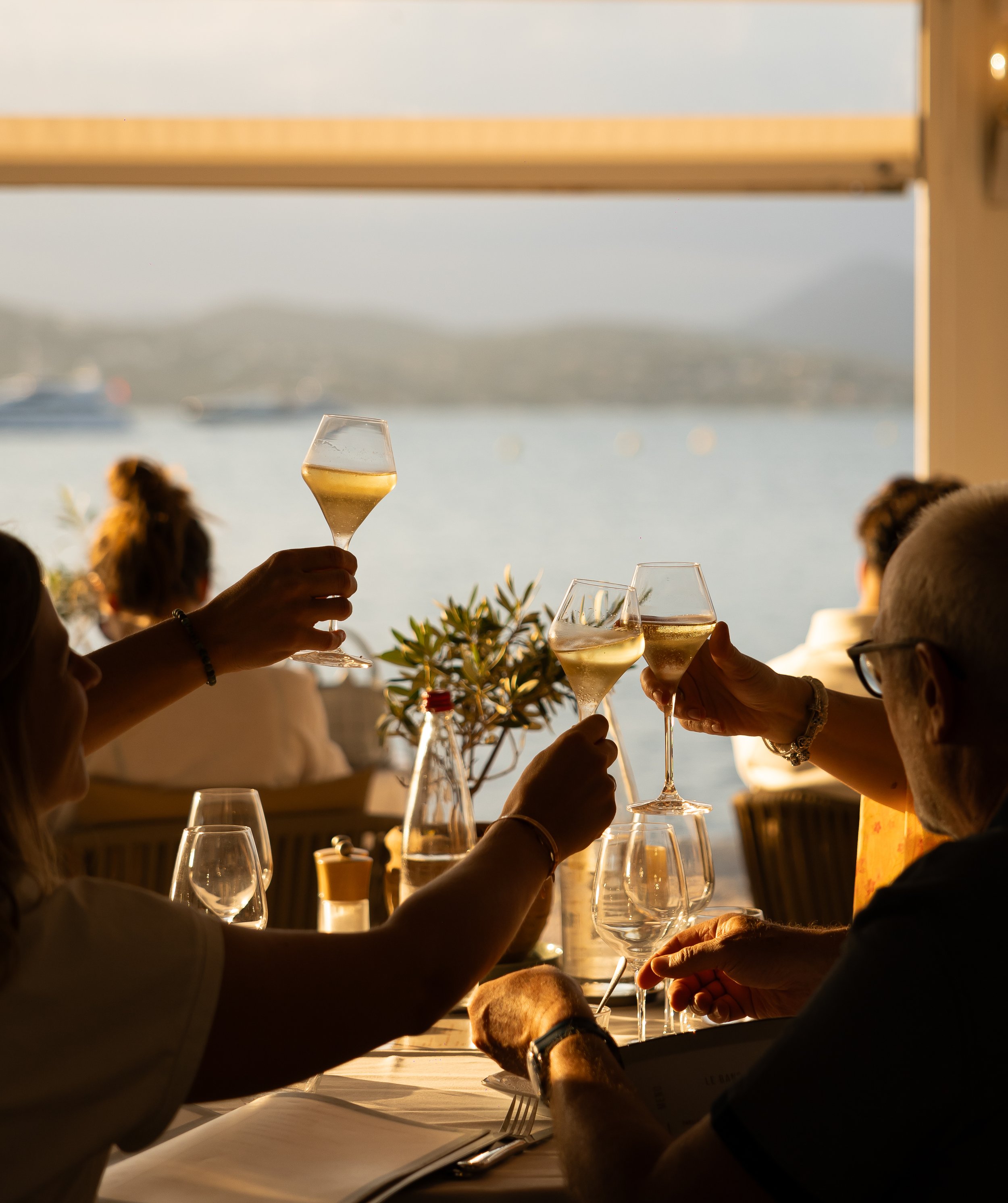 People raising glasses of white wine at a dinner table with a scenic water view during sunset.
