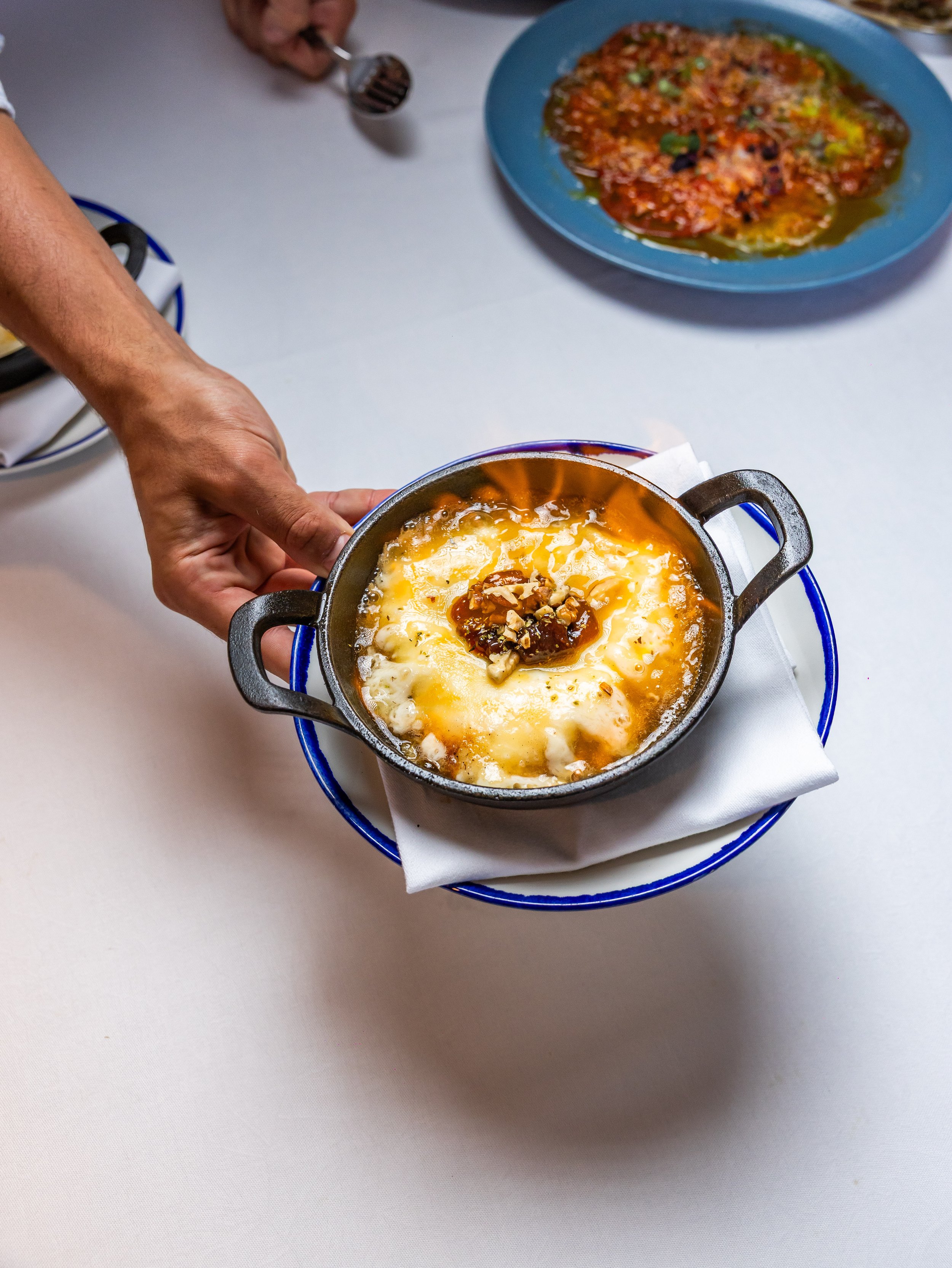 A small black cast-iron skillet with melted cheese, topped with chopped nuts and caramel sauce, on a blue-rimmed plate with a white napkin underneath. In the background, a hand is holding a small spoon over a white surface, and a blue plate with a pa