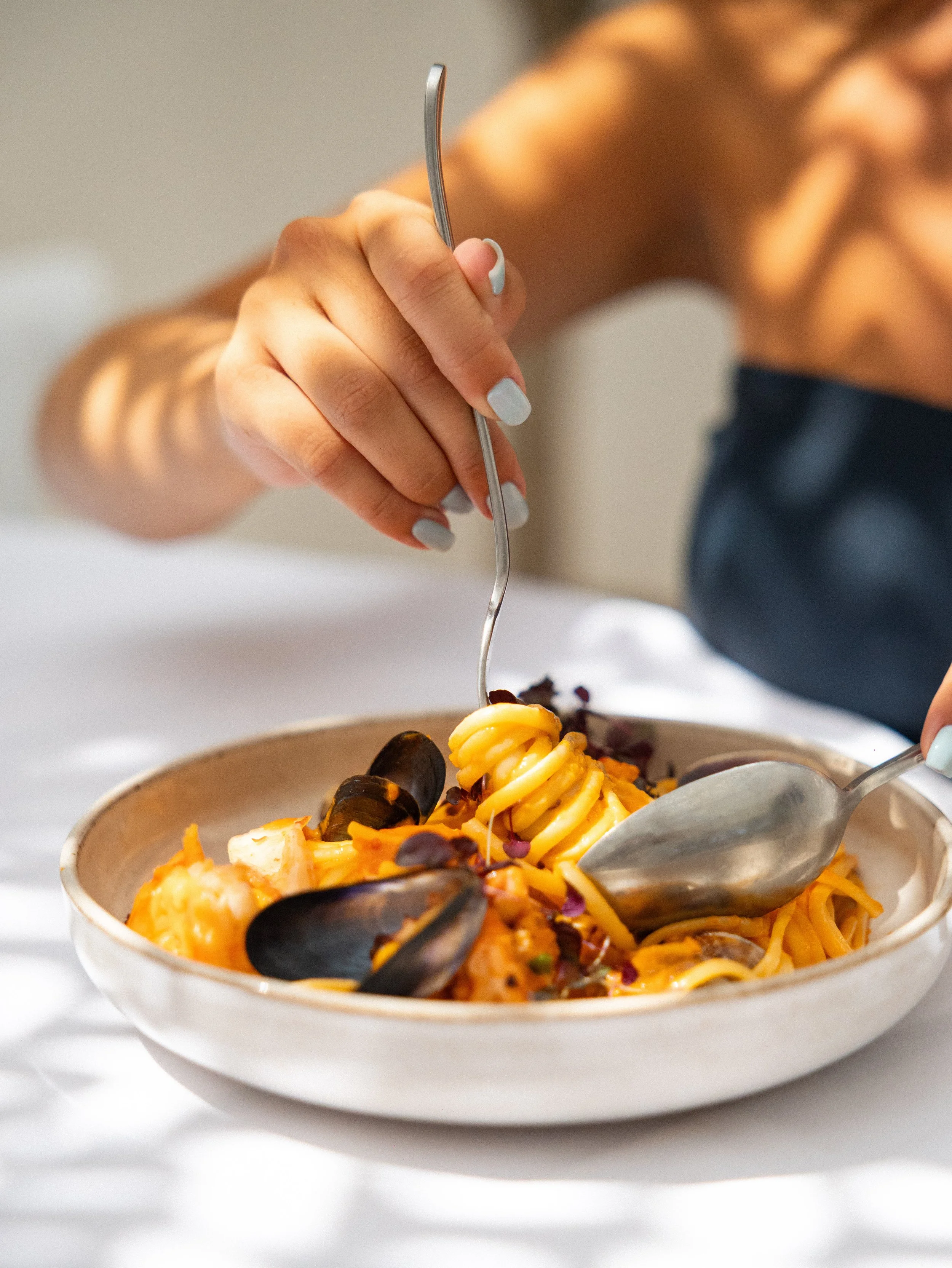 Close-up of a bowl of seafood pasta with mussels, shrimp, and spiral pasta, garnished with herbs, on a white surface. A person is fork-stabbing the pasta.