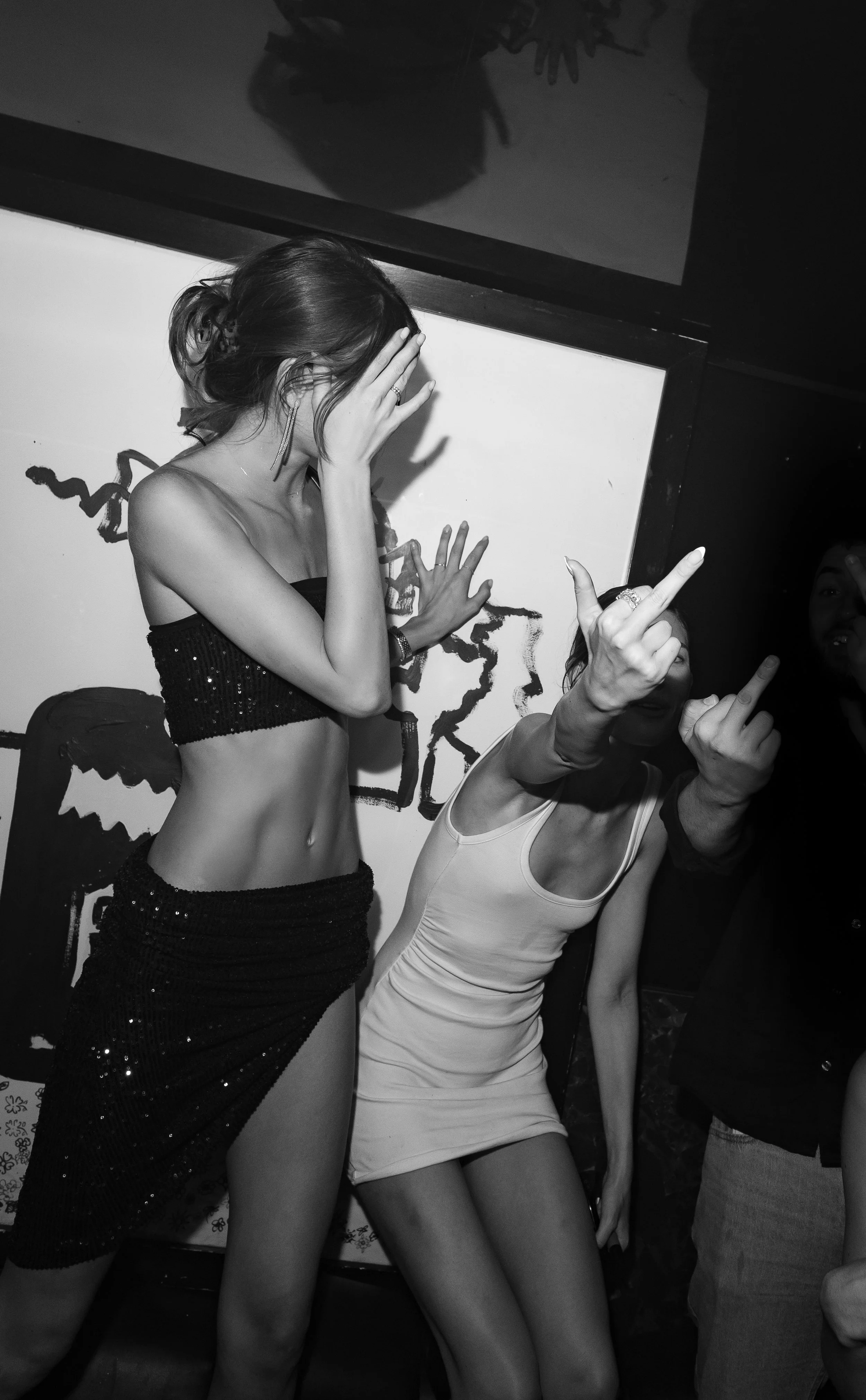 Black and white photo of two women at a party, with one woman covering her face and the other showing her middle finger, their bodies close together.