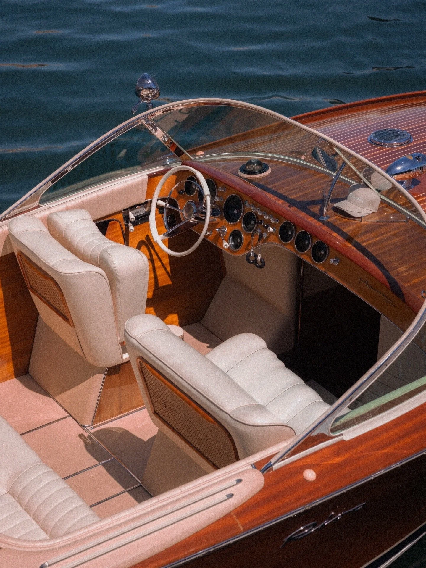 Close-up view of a classic wooden boat's interior with white leather seating, a wooden dashboard with numerous gauges, a white steering wheel, and a beige hat placed on the dashboard, with water visible in the background.