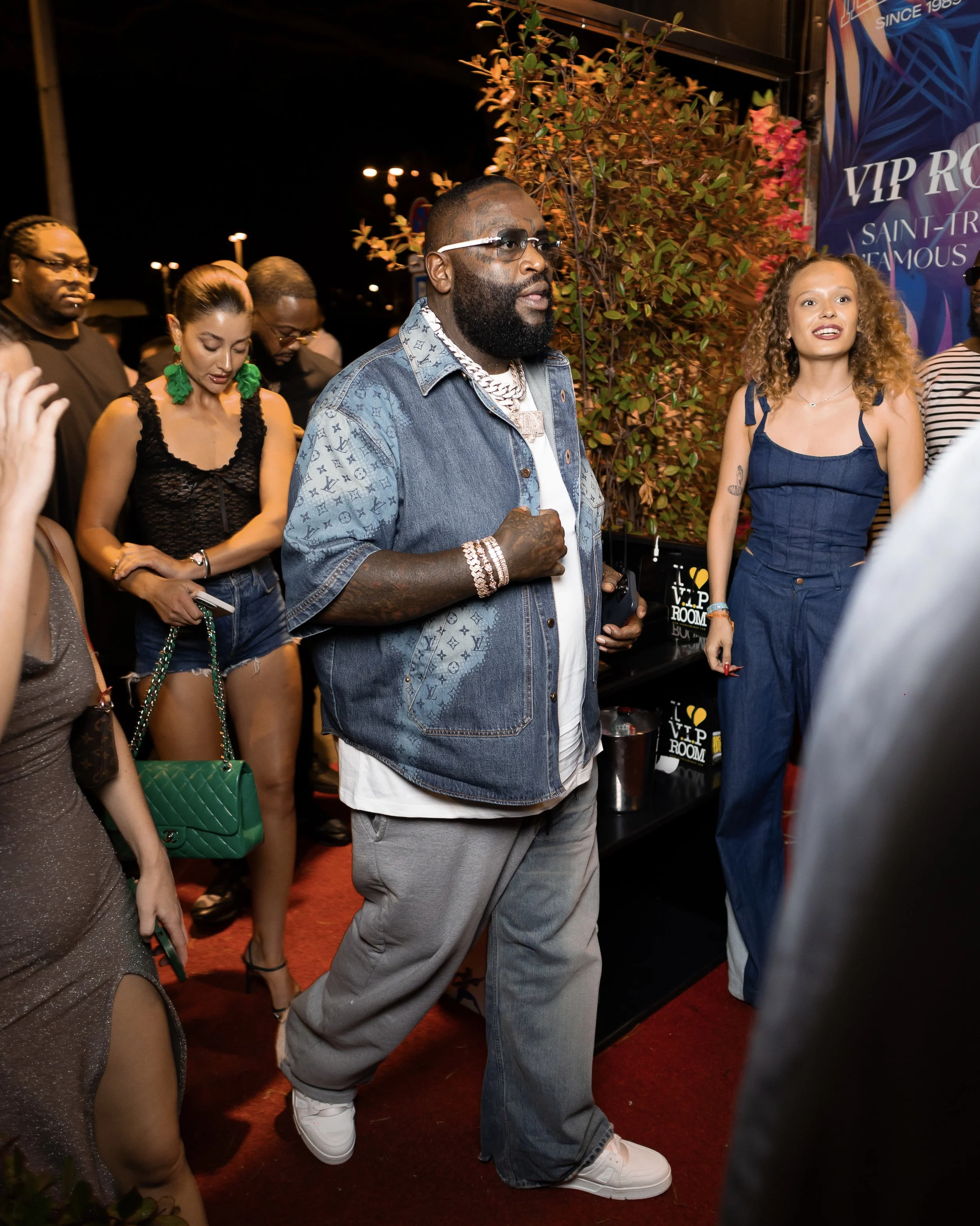 A group of people standing together outdoors at night. The central figure is a man with a beard and sunglasses, wearing a denim jacket, multiple chains, and jewelry. To his right, a woman with curly hair in a blue dress is smiling. Other individuals 