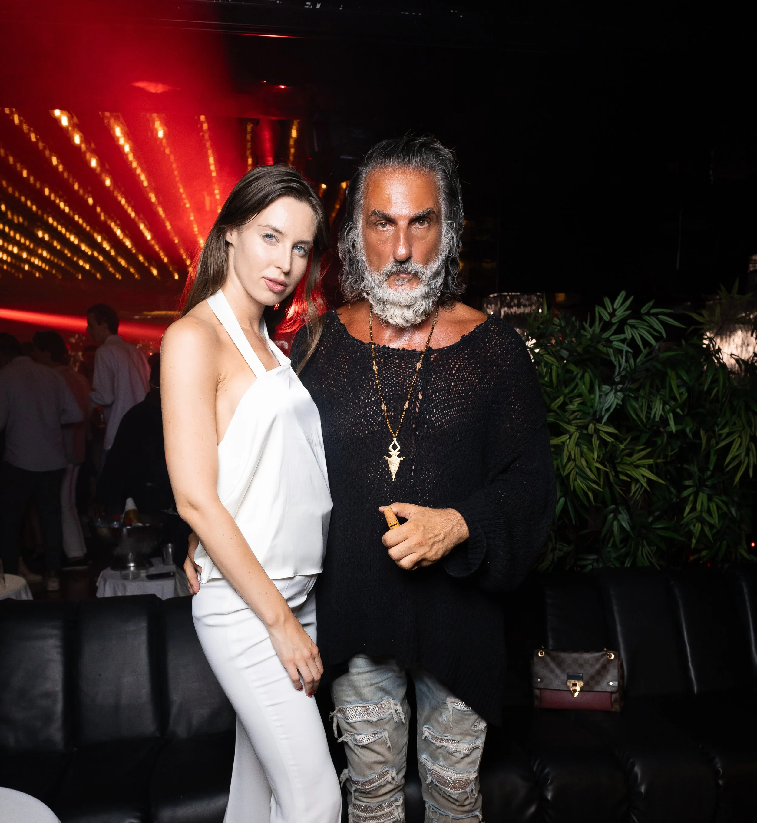 Two people posing together at a party or event. The woman on the left has long brown hair and is wearing a white top or dress. The man on the right has long dark hair and a beard, wearing a black top with a large pendant necklace. The background feat