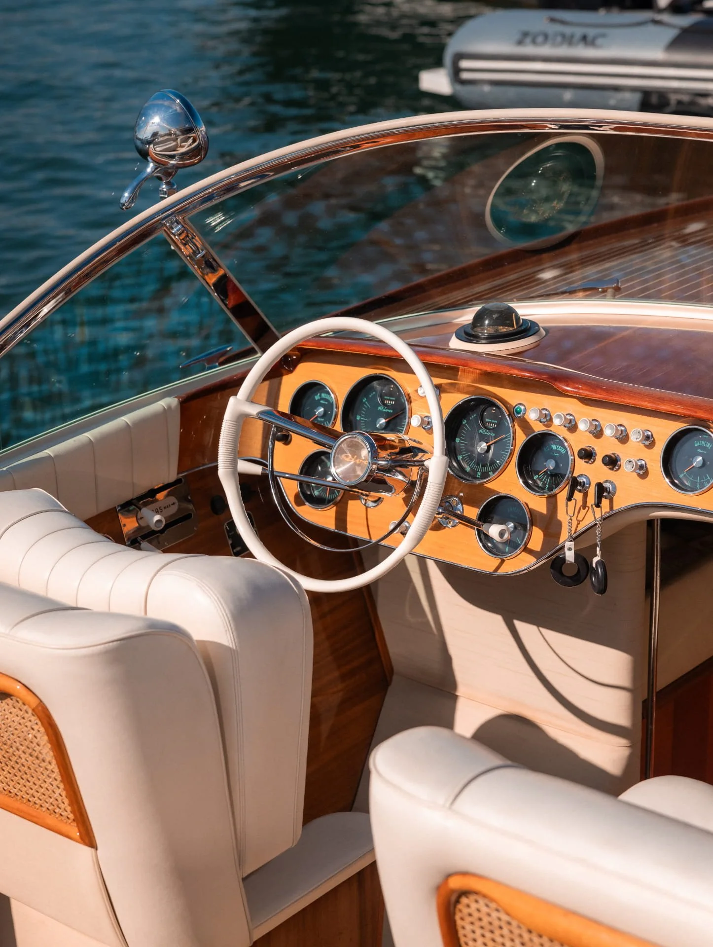 Interior view of a vintage boat with white leather seats, a wooden dashboard with multiple gauges, a white steering wheel, and a chrome rearview mirror, with water visible outside.