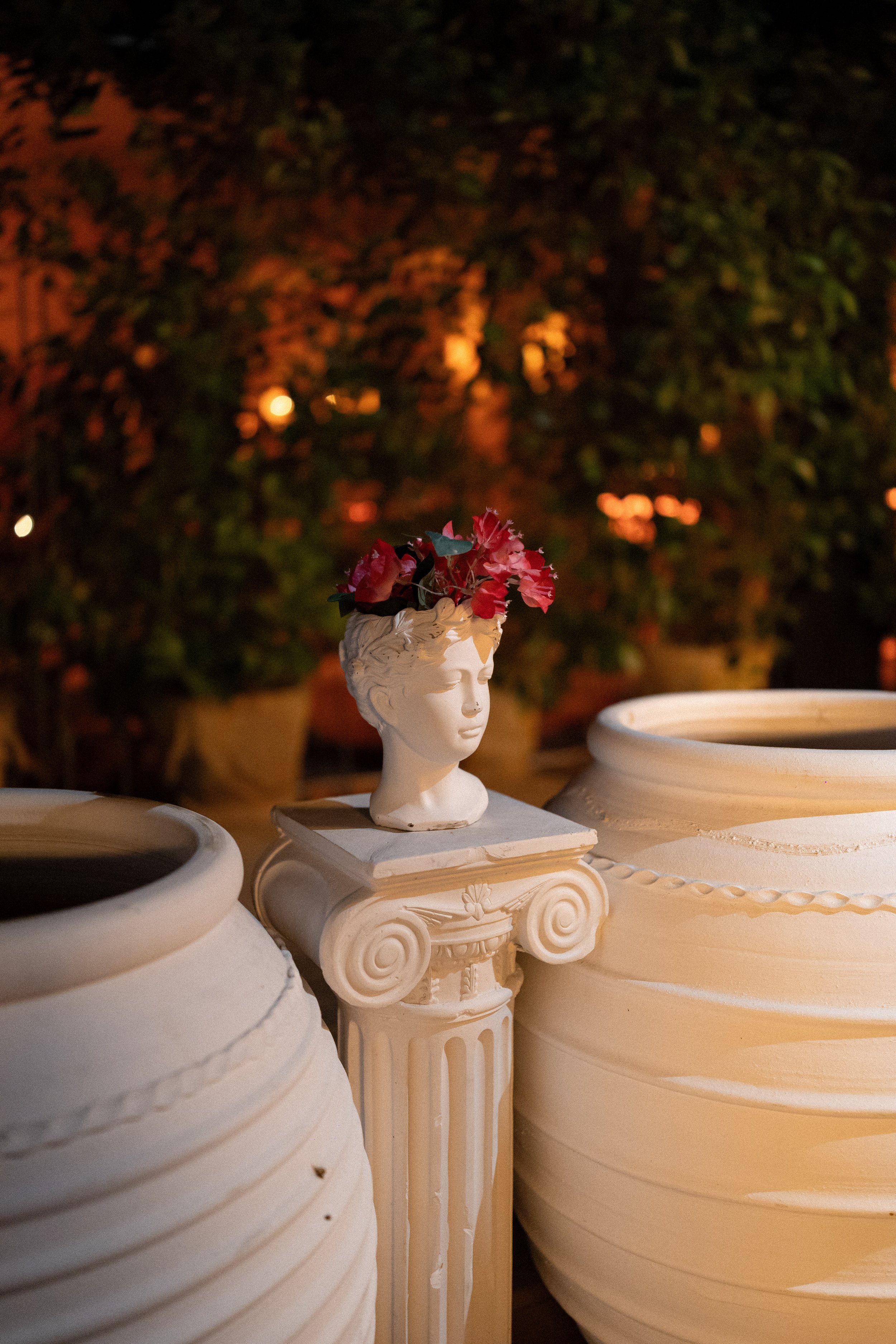 A decorative white bust sculpture of a woman's head with pink flowers in it, placed among large white vases at night with background of blurred trees and warm lighting.