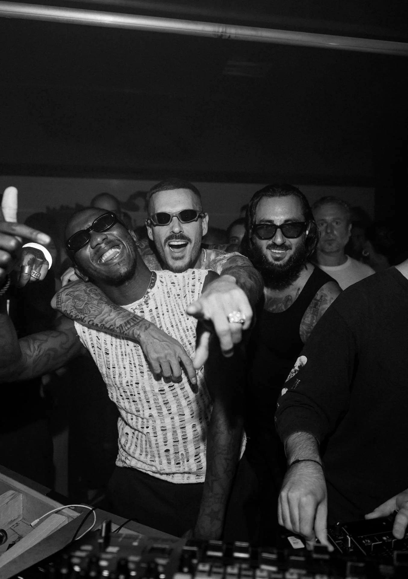 Group of four men at a party or club, wearing sunglasses, smiling, and posing for the camera. One man is pointing, and another is using DJ equipment.