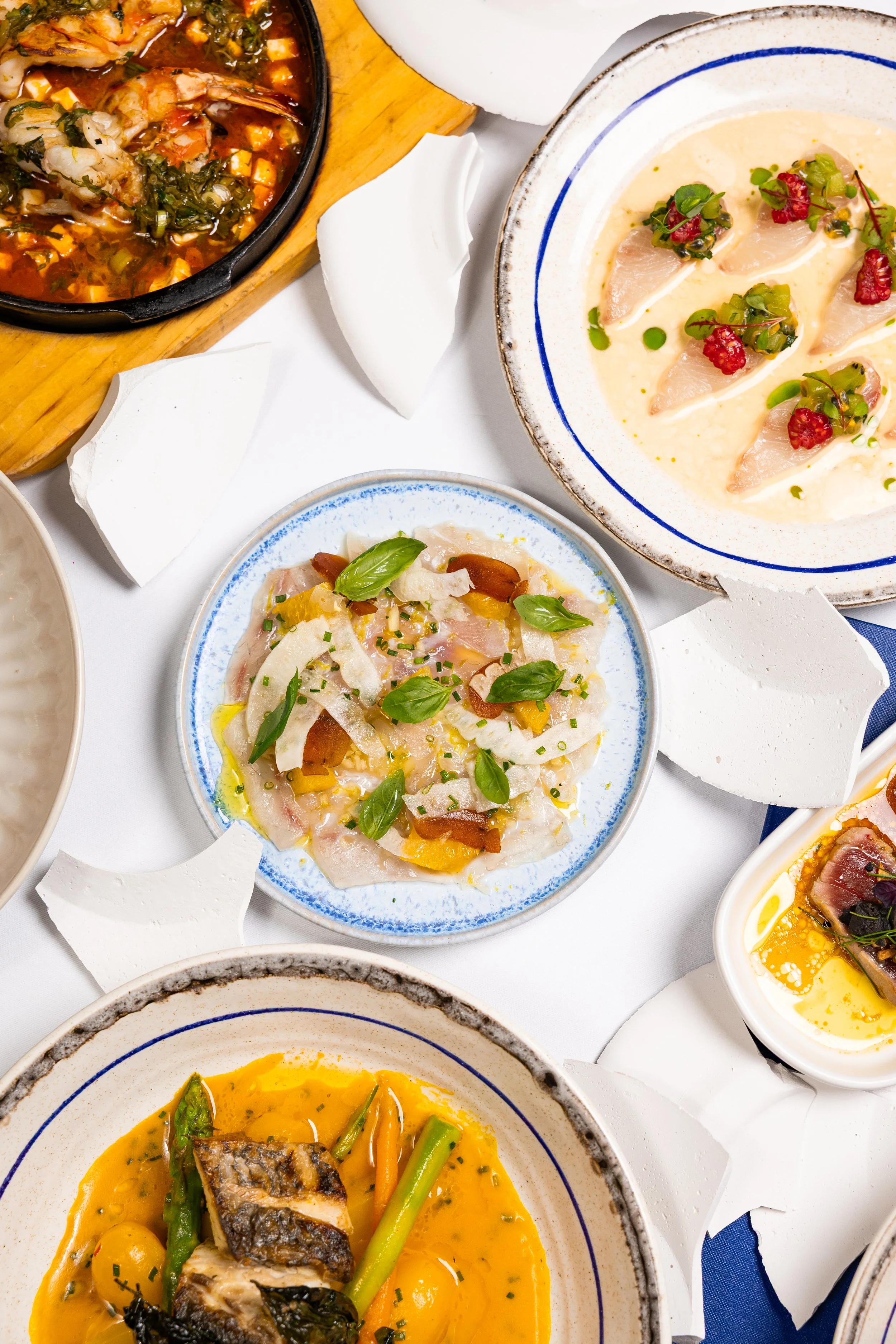 Assorted dishes of seafood, fish, and vegetables served in bowls and plates, including a fish dish with vegetables in a yellow sauce, and a seafood dish with herbs and red berries.