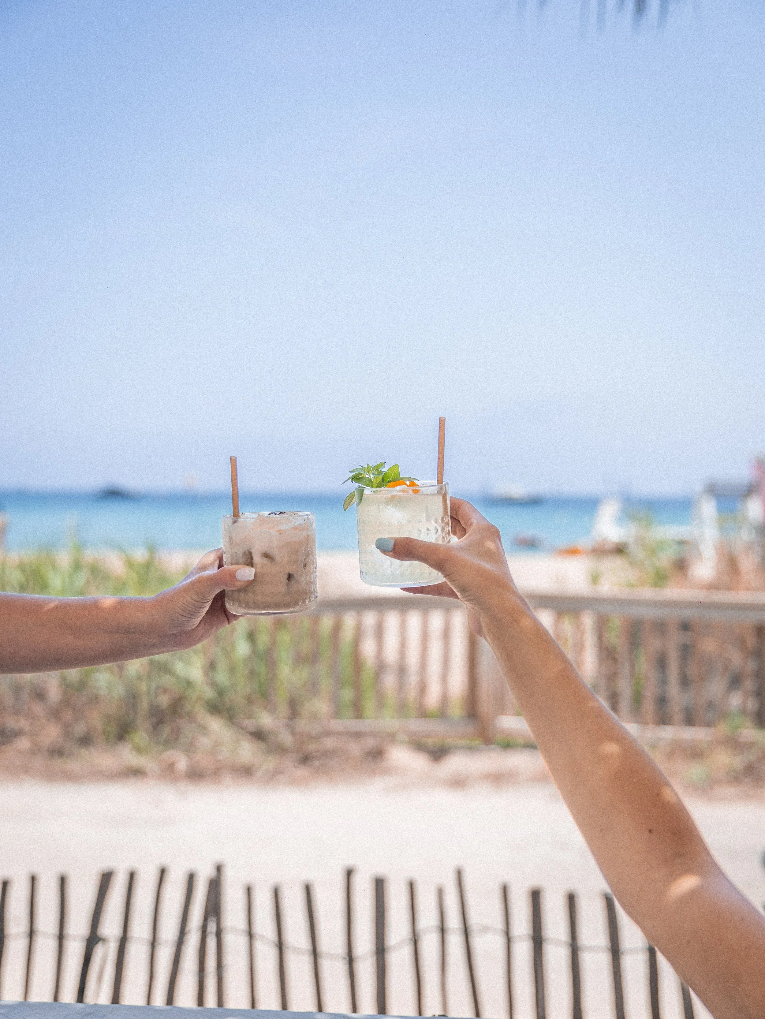 Two hands holding glasses of refreshing drinks with straws and garnishes at a beachside setting with ocean and sky in the background.