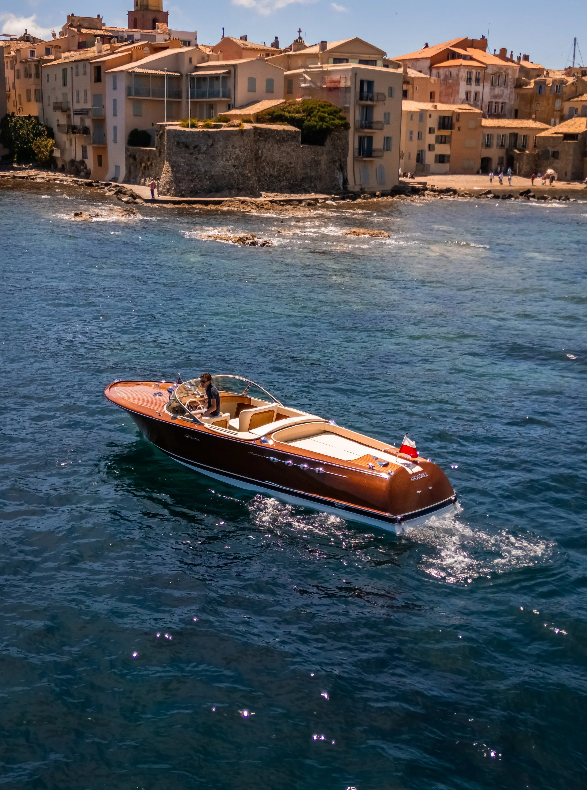 A man sitting in a small motorboat with beige seating and a dark hull, cruising on blue water near a rocky shoreline with buildings and a stone fortress wall in the background.
