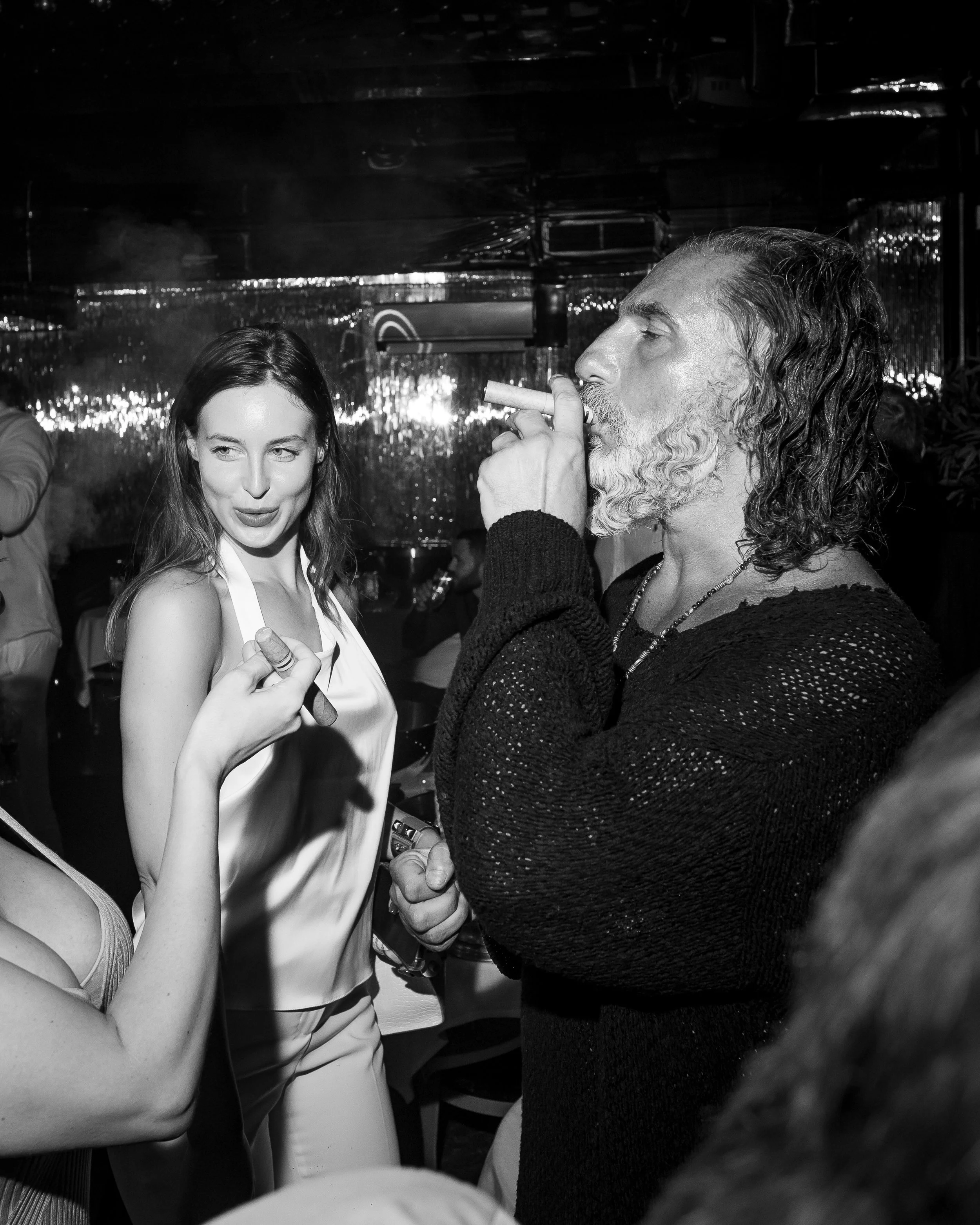 Two people inside a nightclub or bar, one man with long curly hair and a beard smoking a cigar, and a woman with long hair, smiling and holding a cigar, with a reflective shiny wall and dim lighting in the background.