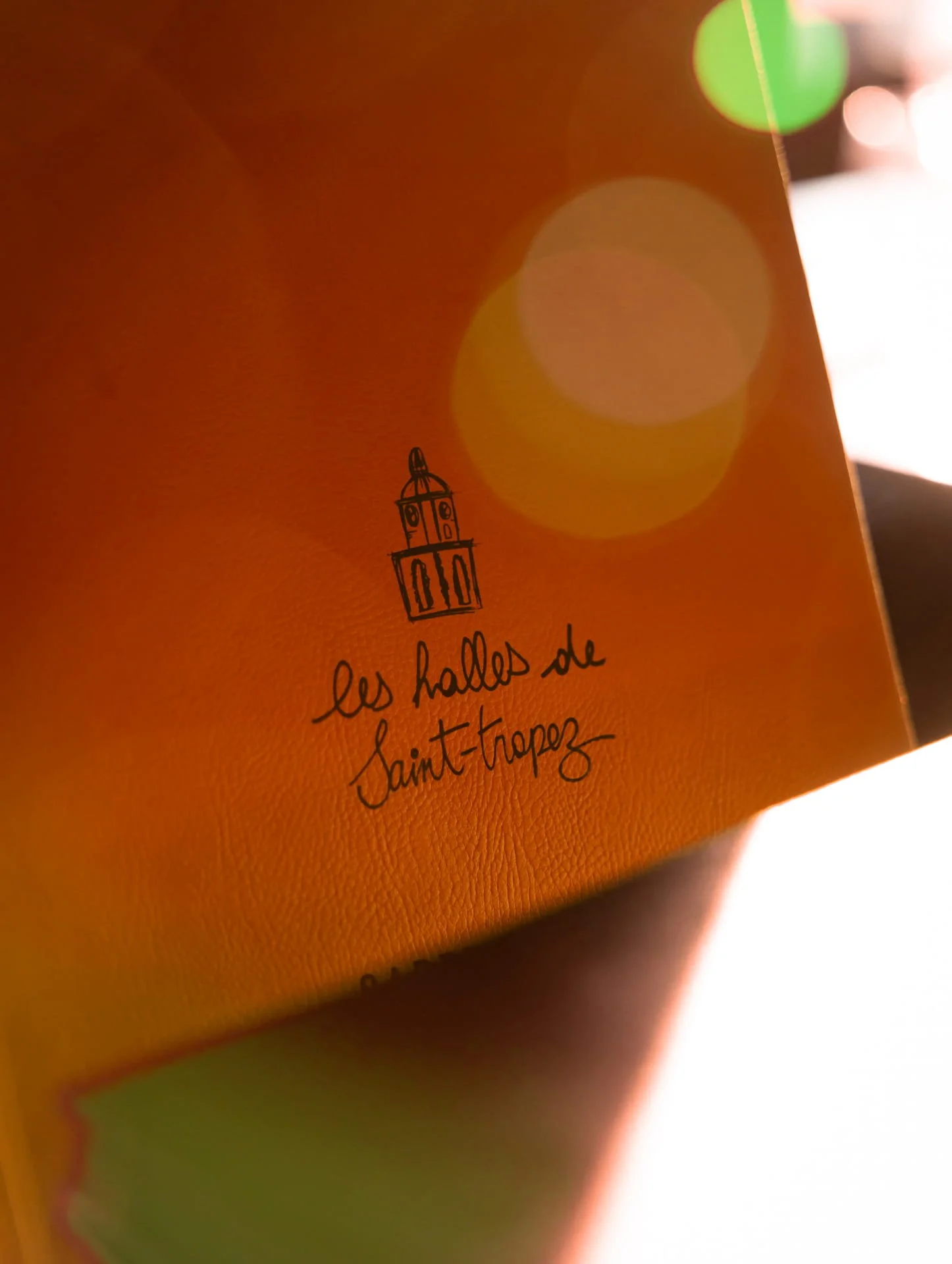 Close-up of a yellow-orange textured paper with a small illustration of a tower and handwritten text that says 'les halles de Saint-Tropez'.