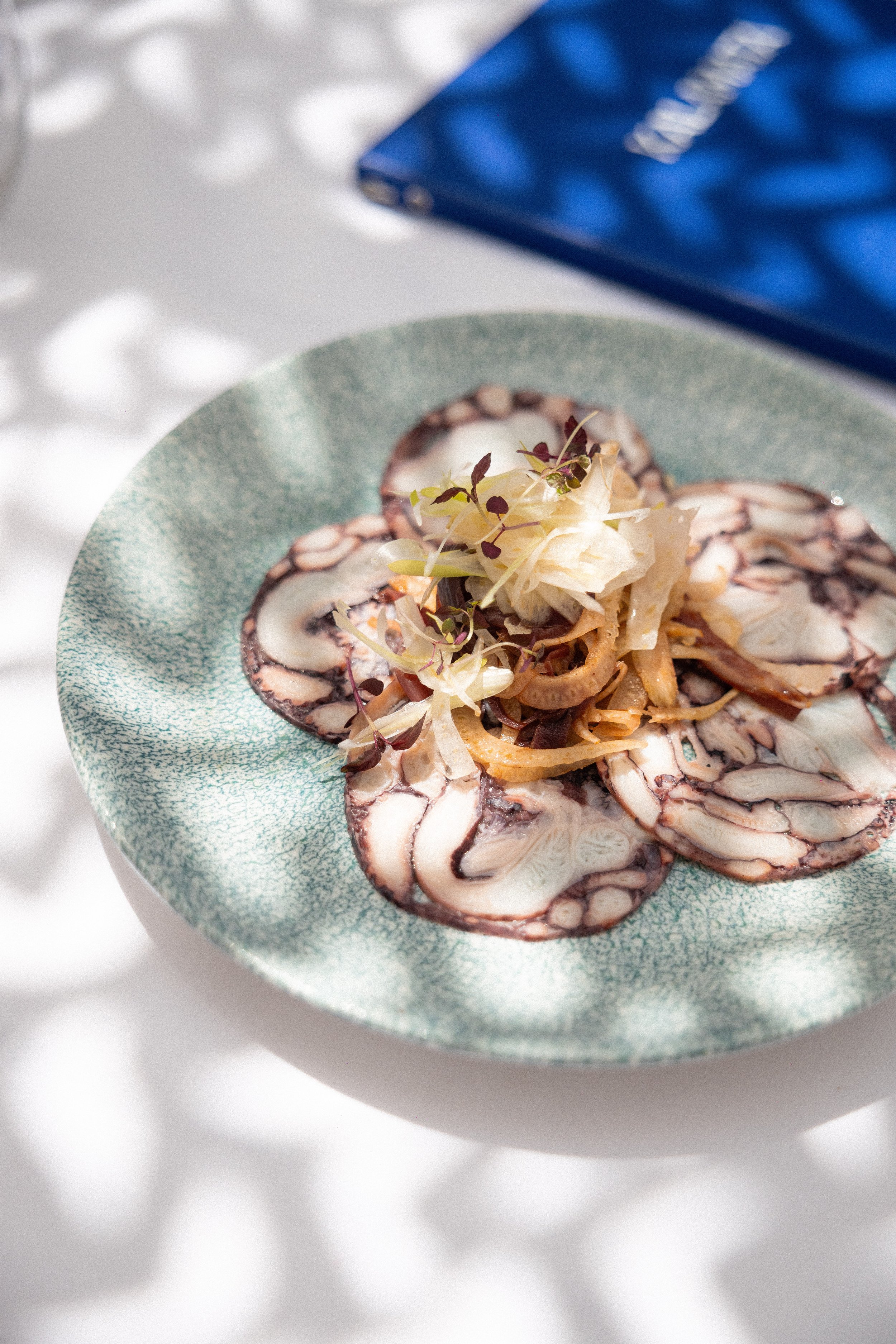 Thin slices of octopus on a green textured plate, garnished with shredded cabbage and microgreens, with a blue book in the background.