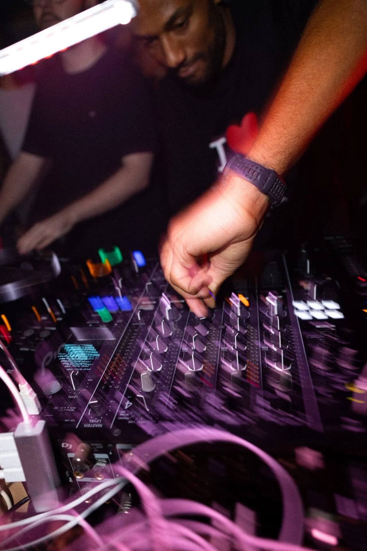 A DJ adjusts controls on a mixing console with his right hand, surrounded by colorful cables and equipment, wearing a black shirt and a purple wristwatch.