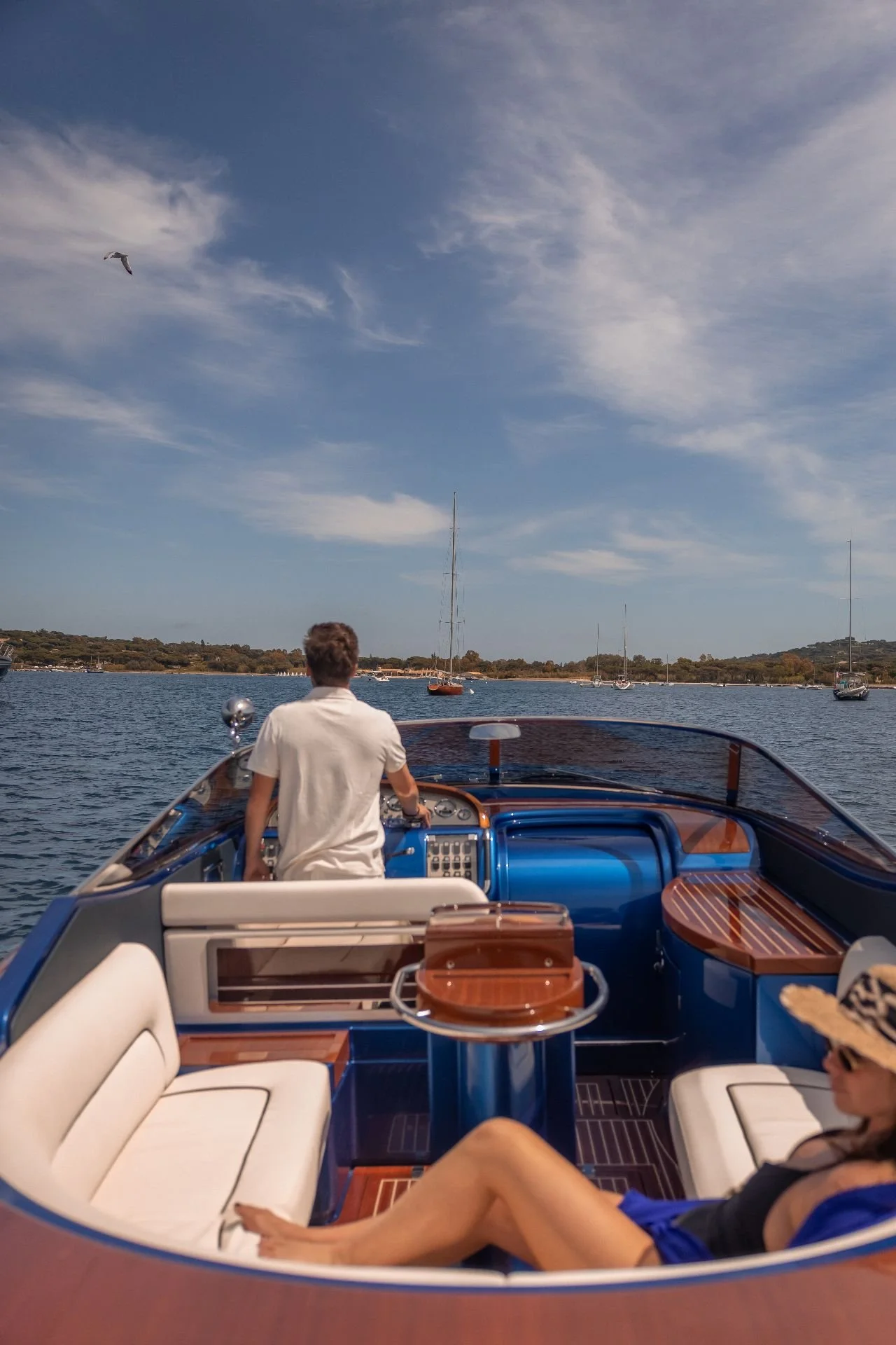 View from a boat showing a man steering, seating area with white cushions, and a person relaxing on the side, with a calm lake, sailboats, and a partly cloudy sky in the background.