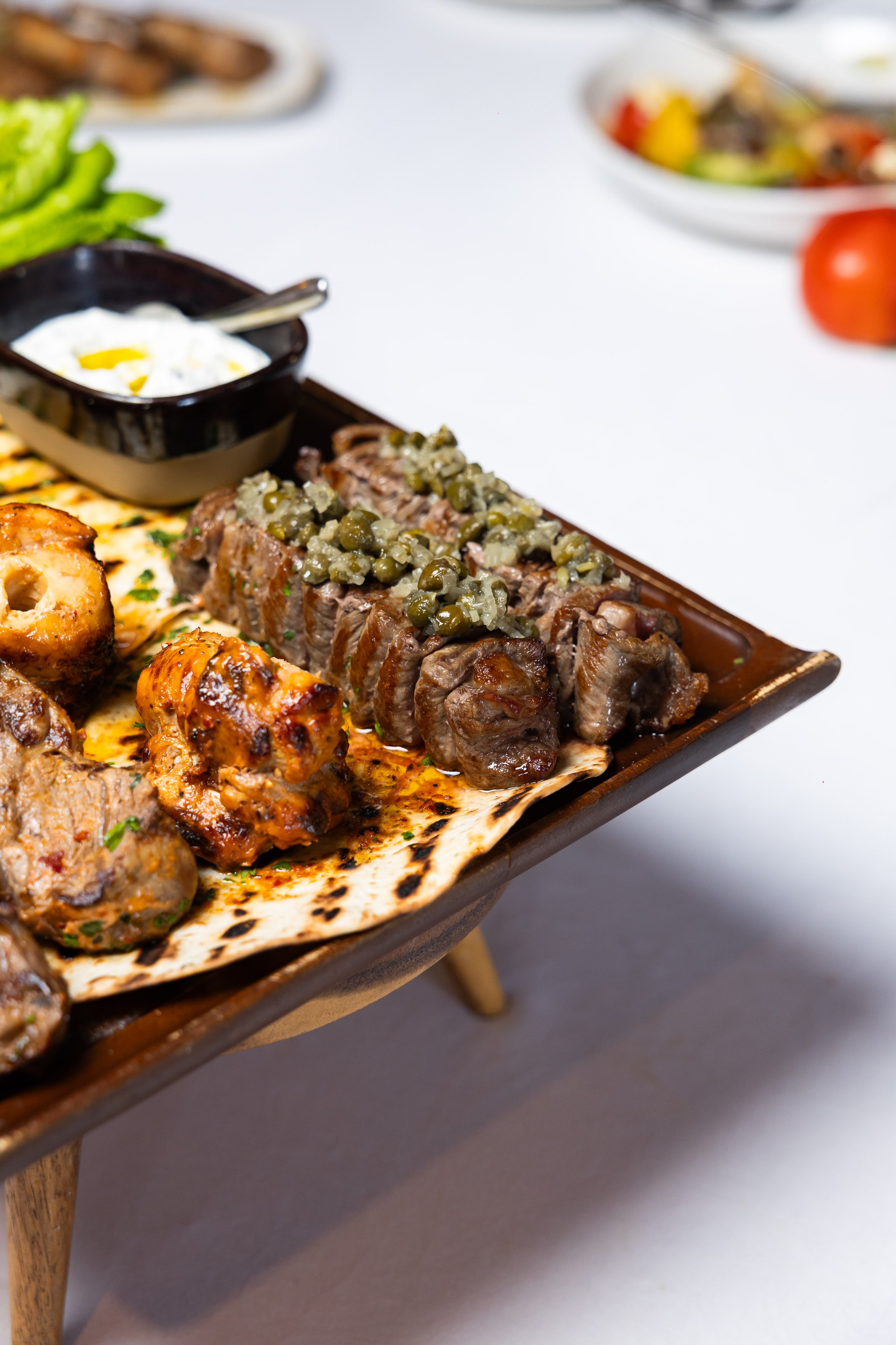 Close-up of a wooden serving platter with grilled meats topped with green peppercorns, a bowl of creamy white dip with a lemon wedge, and grilled vegetables. A small bowl with more sauce, and a plate of meat in the background, with a bowl of salad an