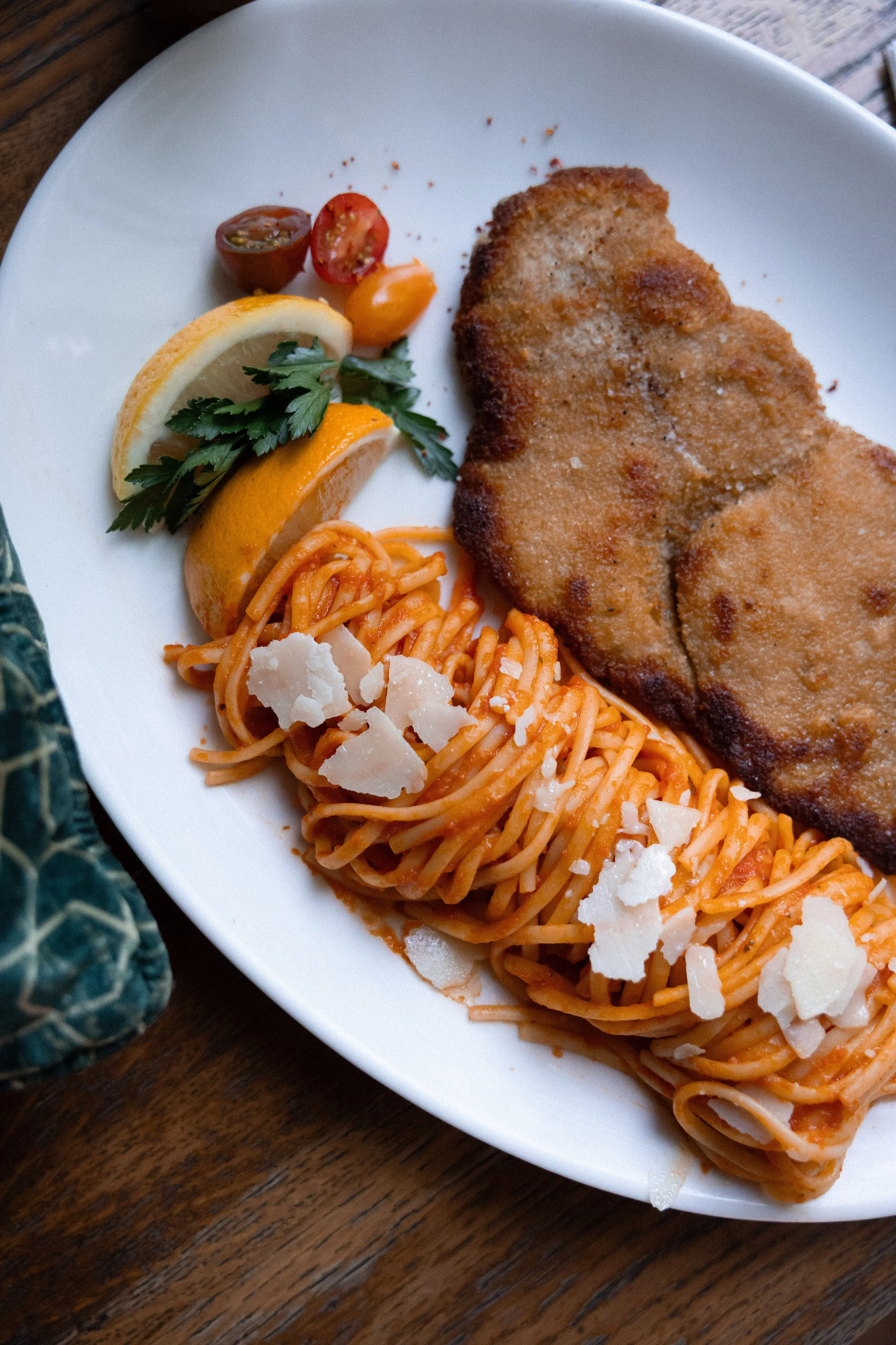 A white plate holding a breaded and fried fish fillet, spaghetti with tomato sauce and grated cheese, cherry tomatoes, lemon slices, and parsley garnish on a wooden table.