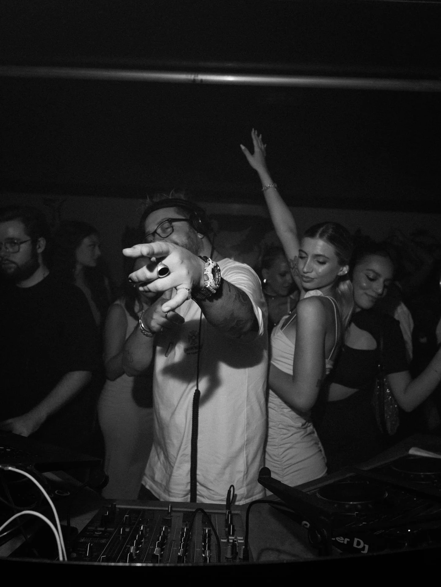 A group of people dancing and having fun at a nightclub. A DJ is in the foreground wearing headphones, pointing, and wearing a watch and rings. Several women are dancing behind him, with one woman raising her arm and others smiling and enjoying the m