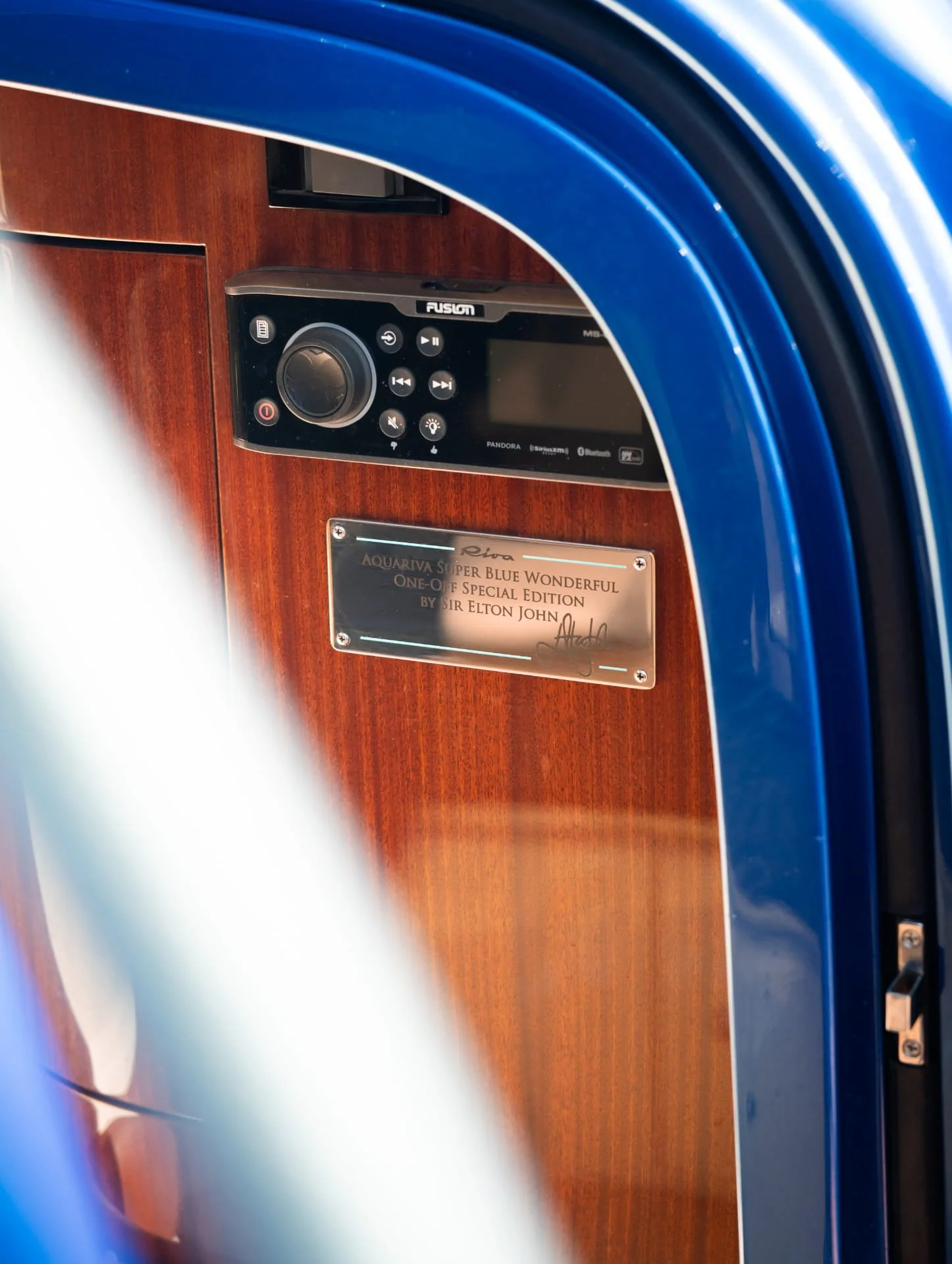 View of a washing machine's control panel and part of its door, with a plaque below it on a wooden surface. The control panel displays various buttons and a large dial, the brand is Fusion, and it includes features like Pandora, SiriusXM, Bluetooth, 