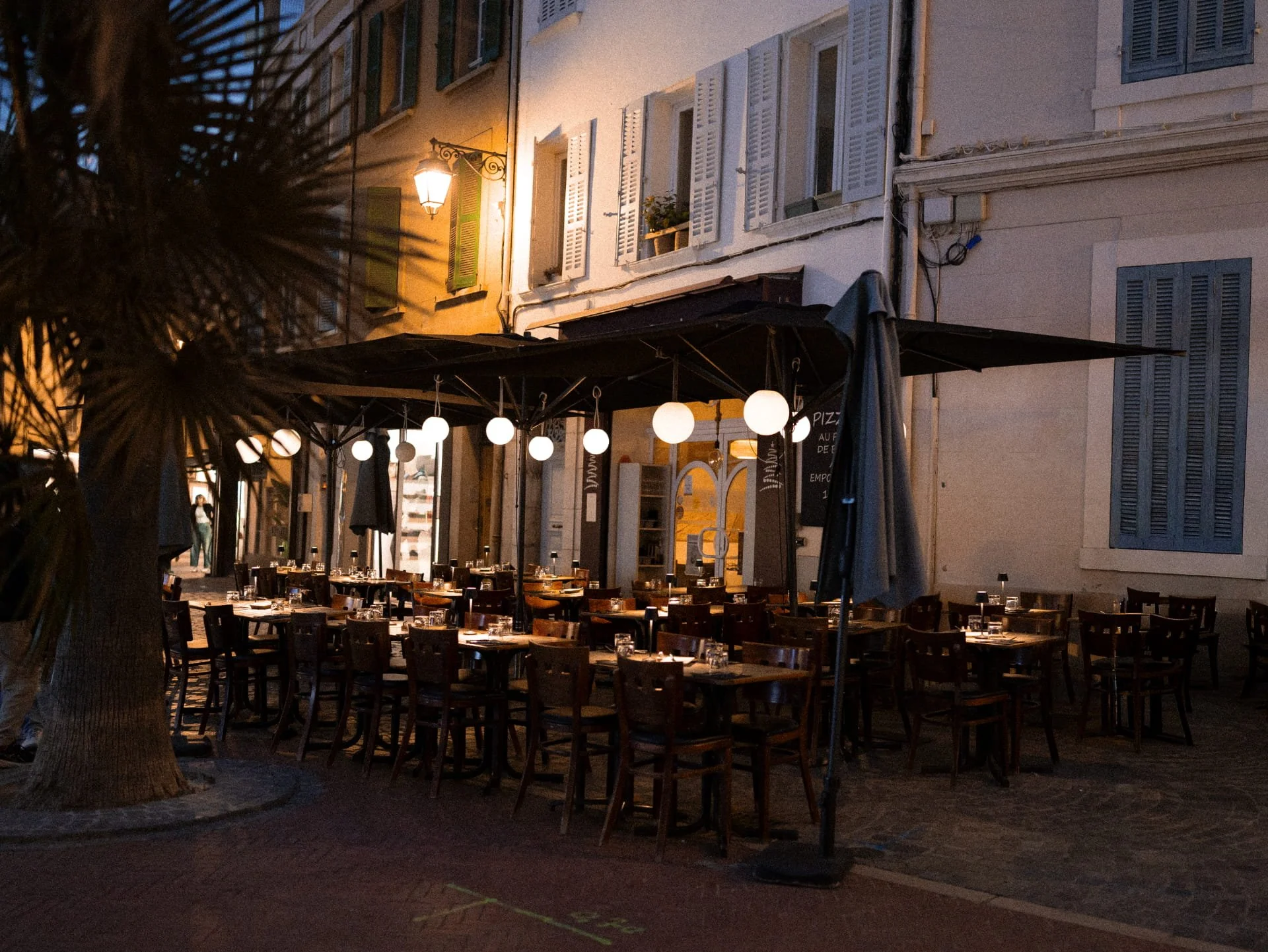 Outdoor restaurant patio with tables and chairs, illuminated by hanging round lights and a streetlamp, in front of residential buildings with shutters and windows at dusk.