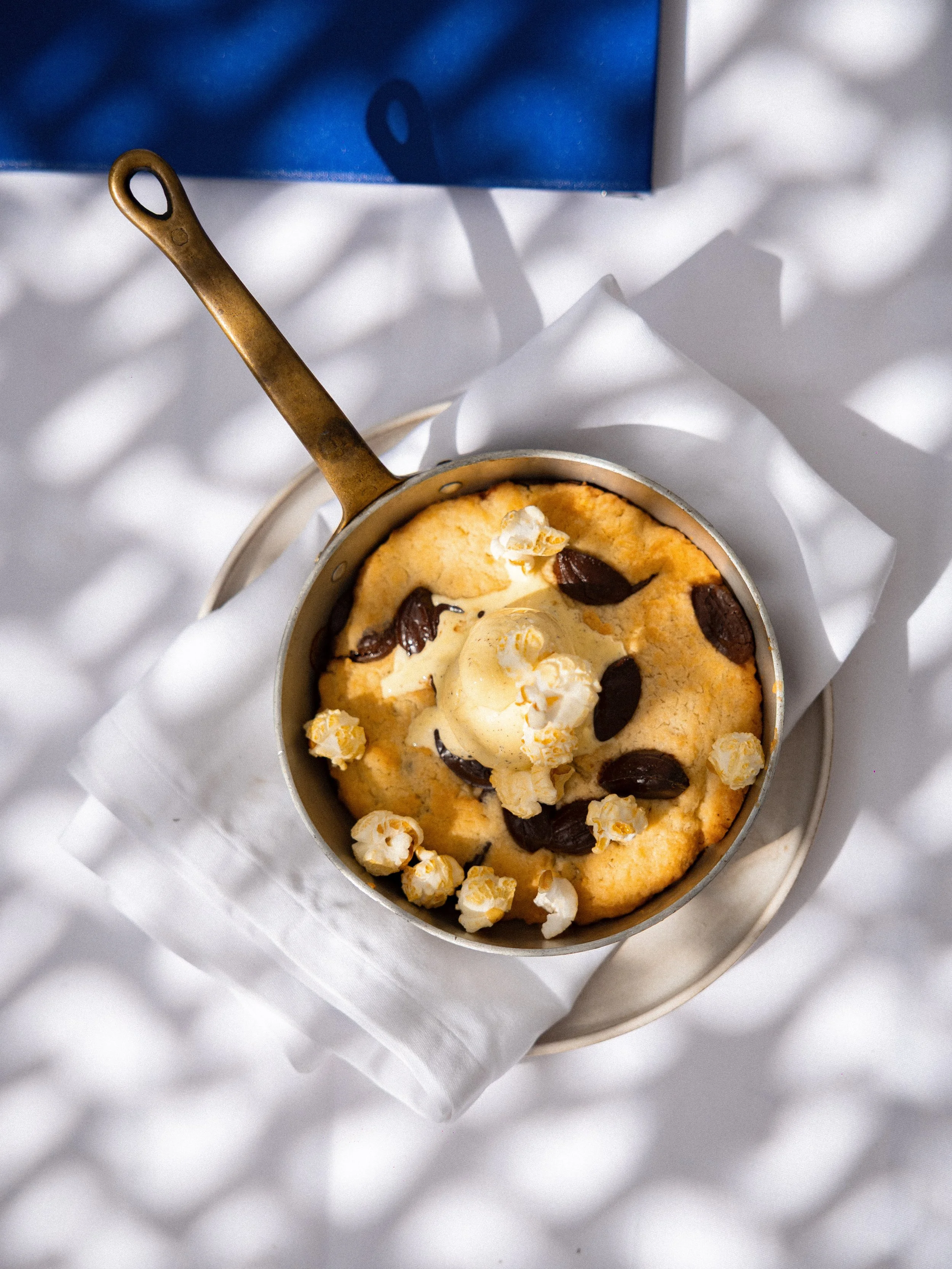 A small round metal container with brownie batter, topped with chocolate chips and popcorn, resting on white crinkled paper on a white surface, with a blue box partially visible in the background.