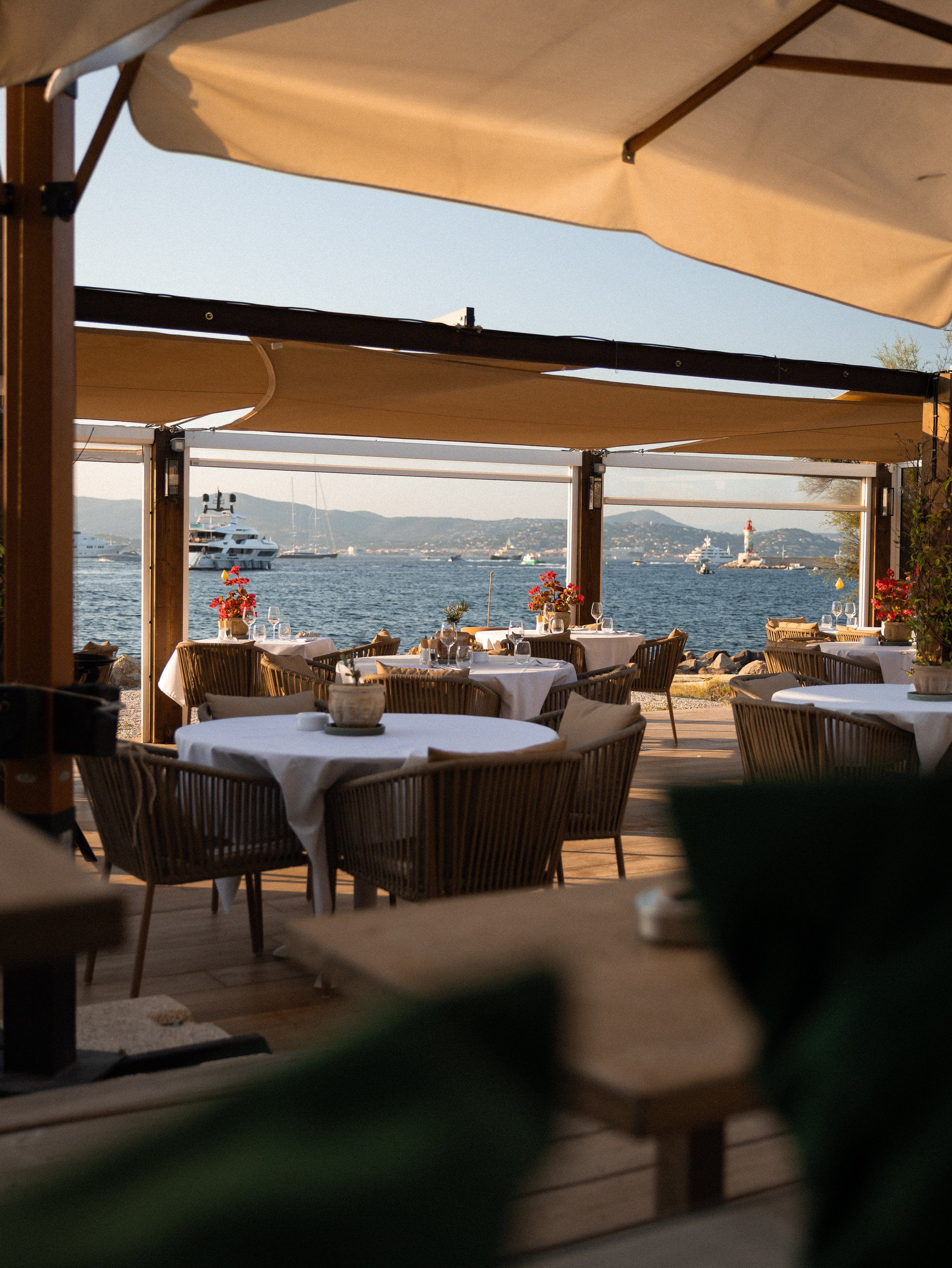 A view of a waterfront restaurant or café with tables set with white tablecloths, glassware, and flowers. The scene is seen through a frame of wooden beams and fabric awnings, overlooking boats and yachts on the water, with mountains and a lighthouse