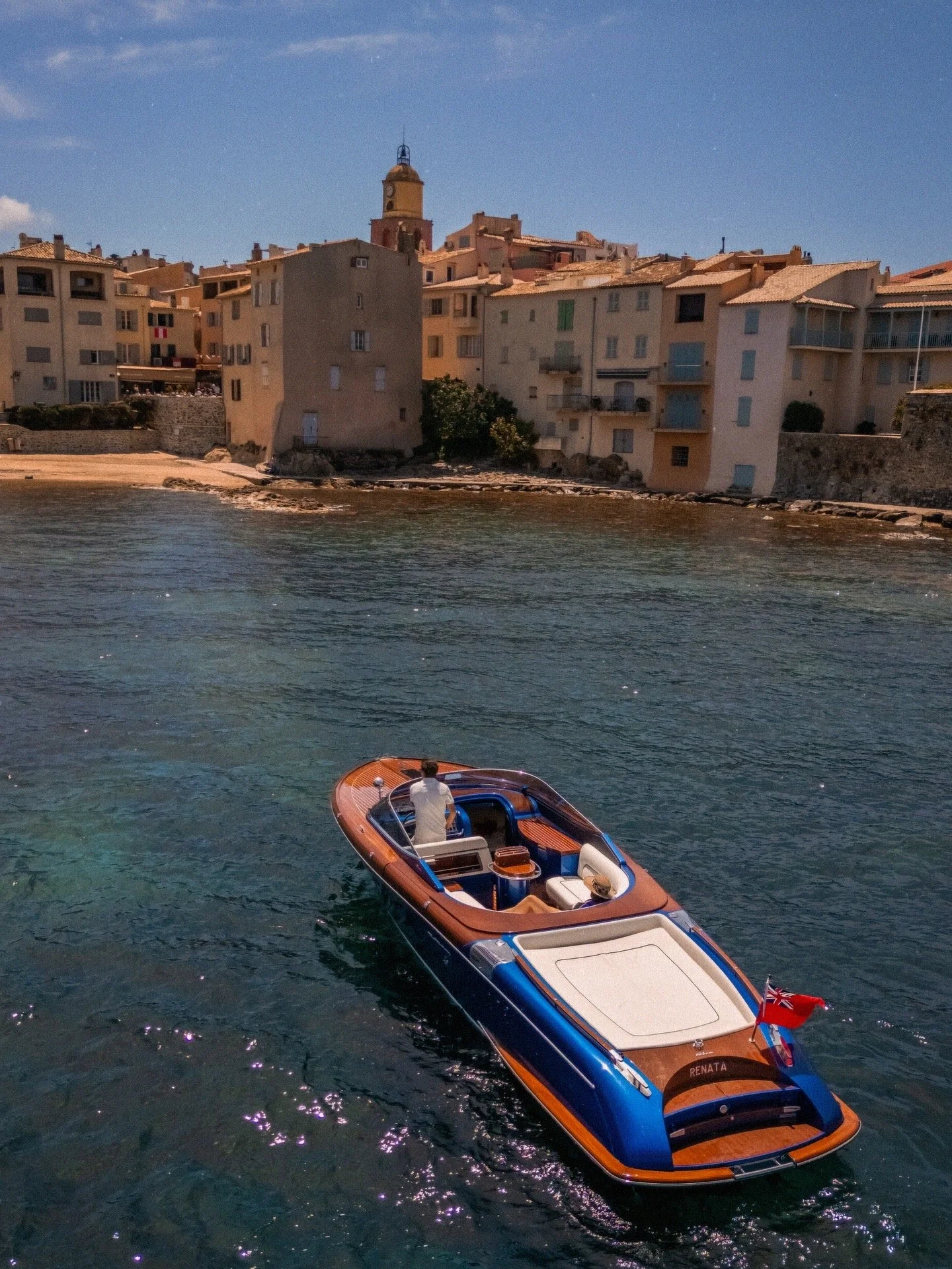 A luxury boat named Renata floating on the water near a coastal town with multi-story buildings and a church tower under a blue sky.