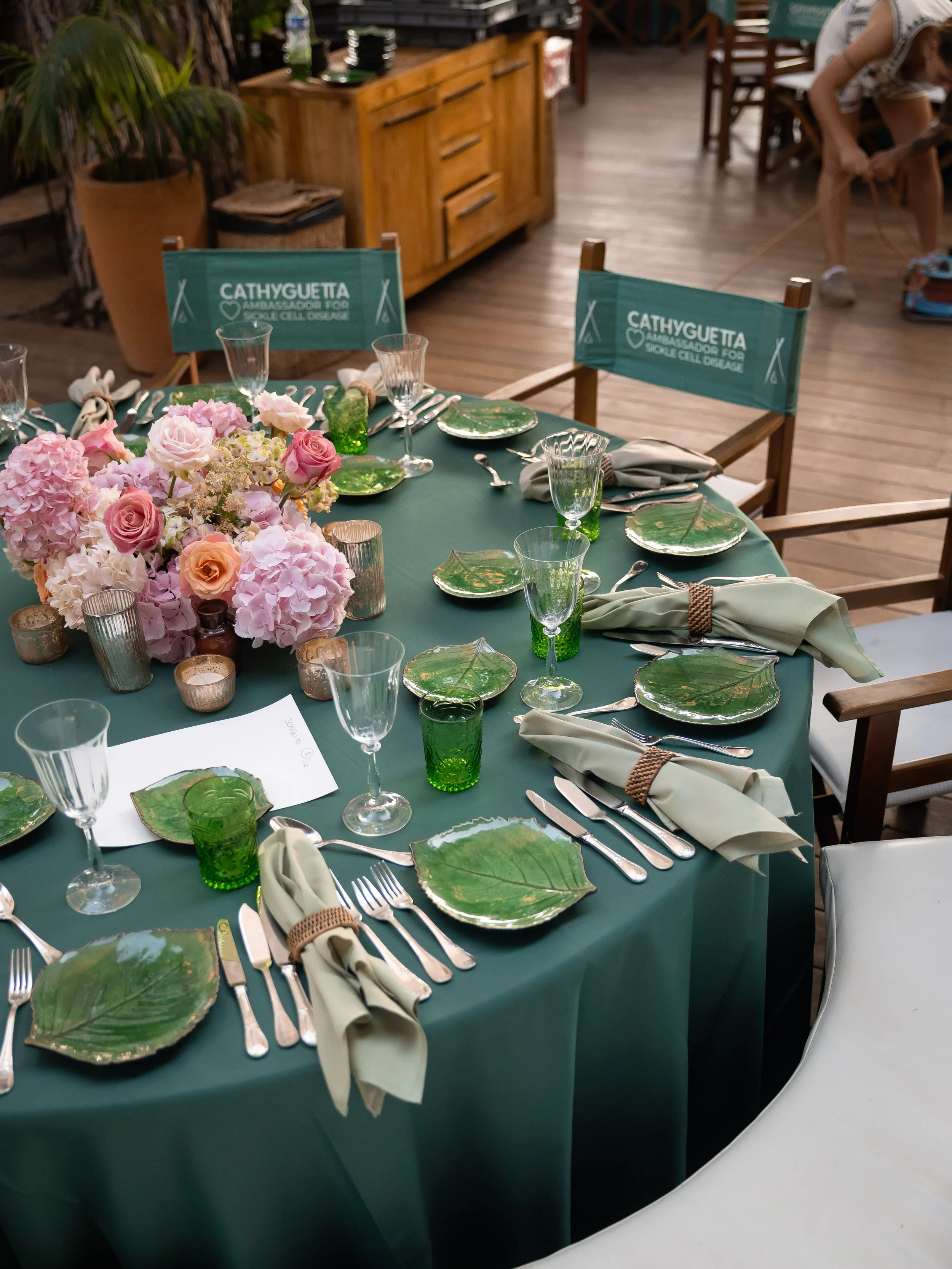 A table set for a meal with pink and peach flowers and green leaf-shaped plates, surrounded by chairs with teal fabric backings that read 'CATHYGUETTA AMBASSADOR FOR SOUL CELL DAMAGE', in a rustic setting with wooden furniture and a person in the bac